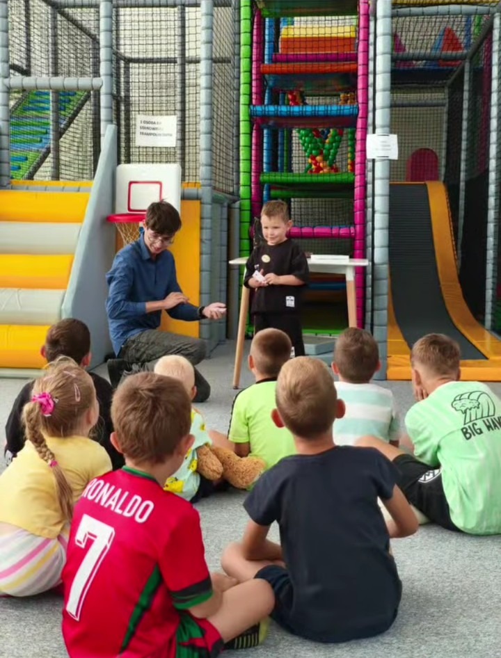 Animator w okularach prowadzi interaktywne zajęcia dla dzieci w sali zabaw z kolorową konstrukcją, gdzie dzieci siedzą na podłodze, słuchając z zaciekawieniem.