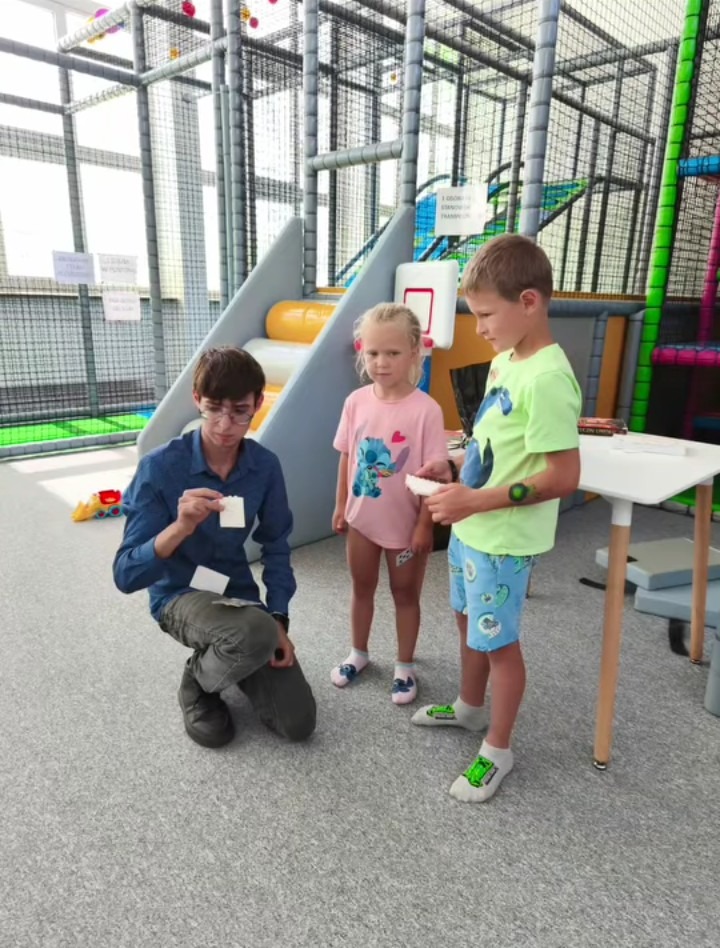 Iluzjonista prezentuje triki karciane dwójce dzieci w sali zabaw. Mężczyzna w okularach klęczy, trzymając karty, a dzieci obserwują z zaciekawieniem. W tle konstrukcja placu zabaw.