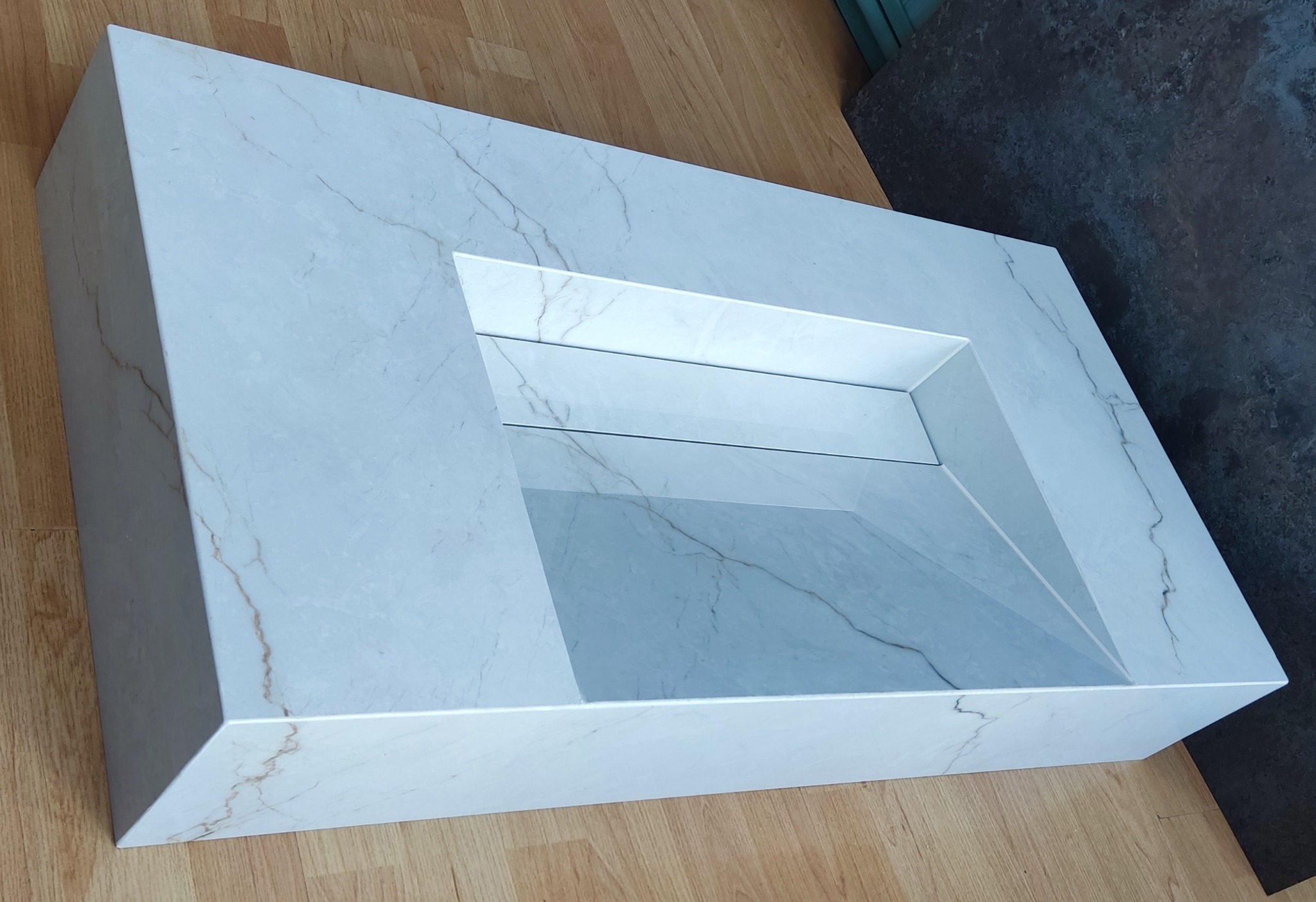 Nowoczesna umywalka z białego marmuru z subtelnym żyłkowaniem, osadzona na drewnianej podłodze. Minimalistyczny design, gładkie powierzchnie i geometryczny kształt.