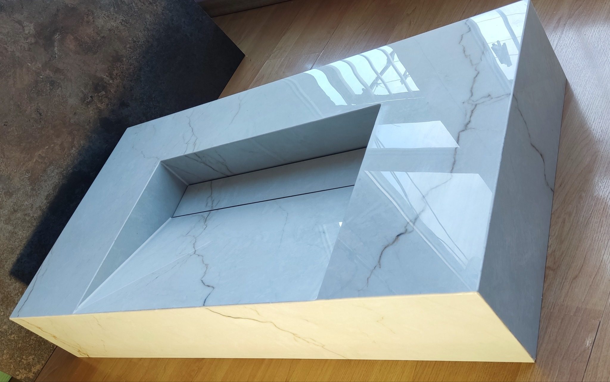 Biała, nowoczesna umywalka z marmurowym wzorem, wykonana z płytek, ustawiona na drewnianej podłodze, z widocznym fragmentem ciemnej płytki w tle.