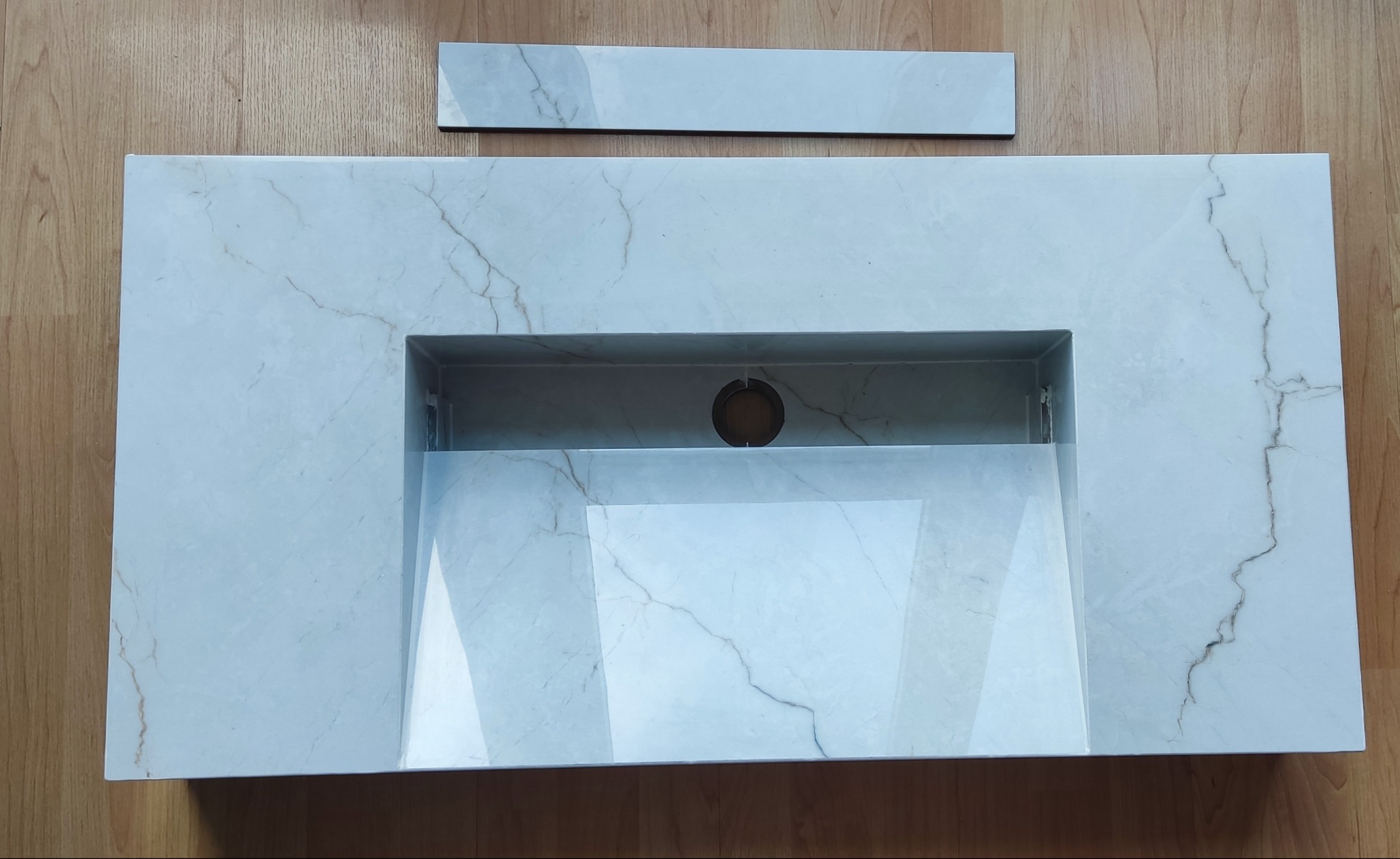 Biała umywalka z marmurowym wzorem i otworem na baterię, leżąca na drewnianej podłodze, z dodatkową płytką obok. Widok z góry.