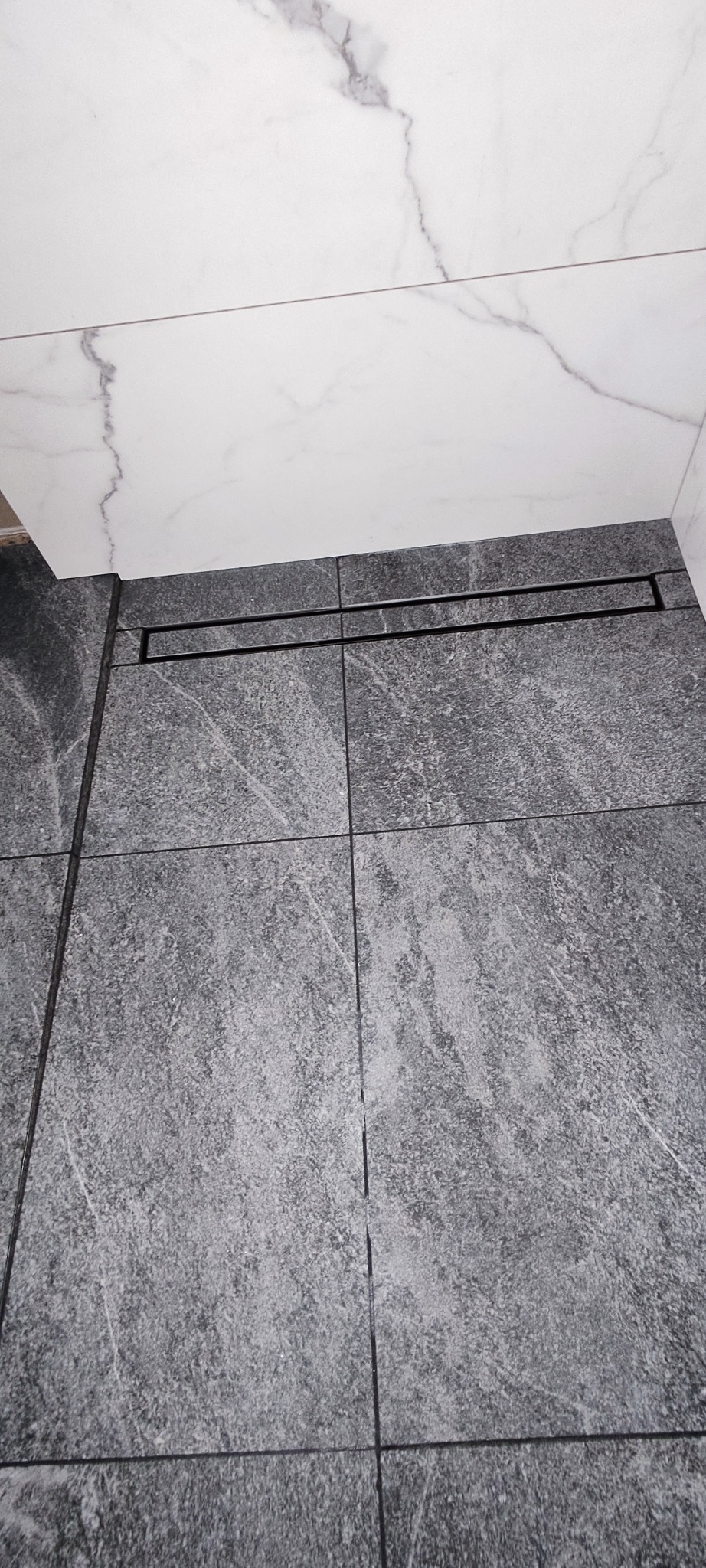 Szare płytki podłogowe z odpływem liniowym i białe płytki ścienne imitujące marmur. Minimalistyczny design łazienki.