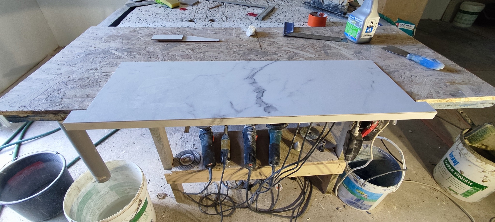 Przygotowany do cięcia marmurowy blat na stole warsztatowym. Widoczne szlifierki kątowe, wiadro z zaprawą i narzędzia glazurnicze. Blat imituje marmur.