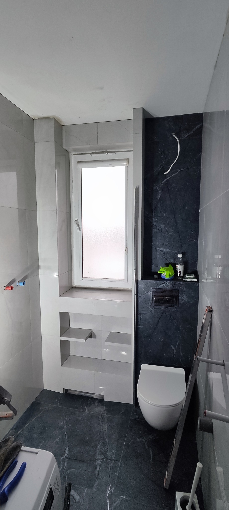 Wykończona łazienka z szarymi płytkami imitującymi marmur, zabudową okna z półkami i wiszącą toaletą. Widoczne narzędzia glazurnicze.
