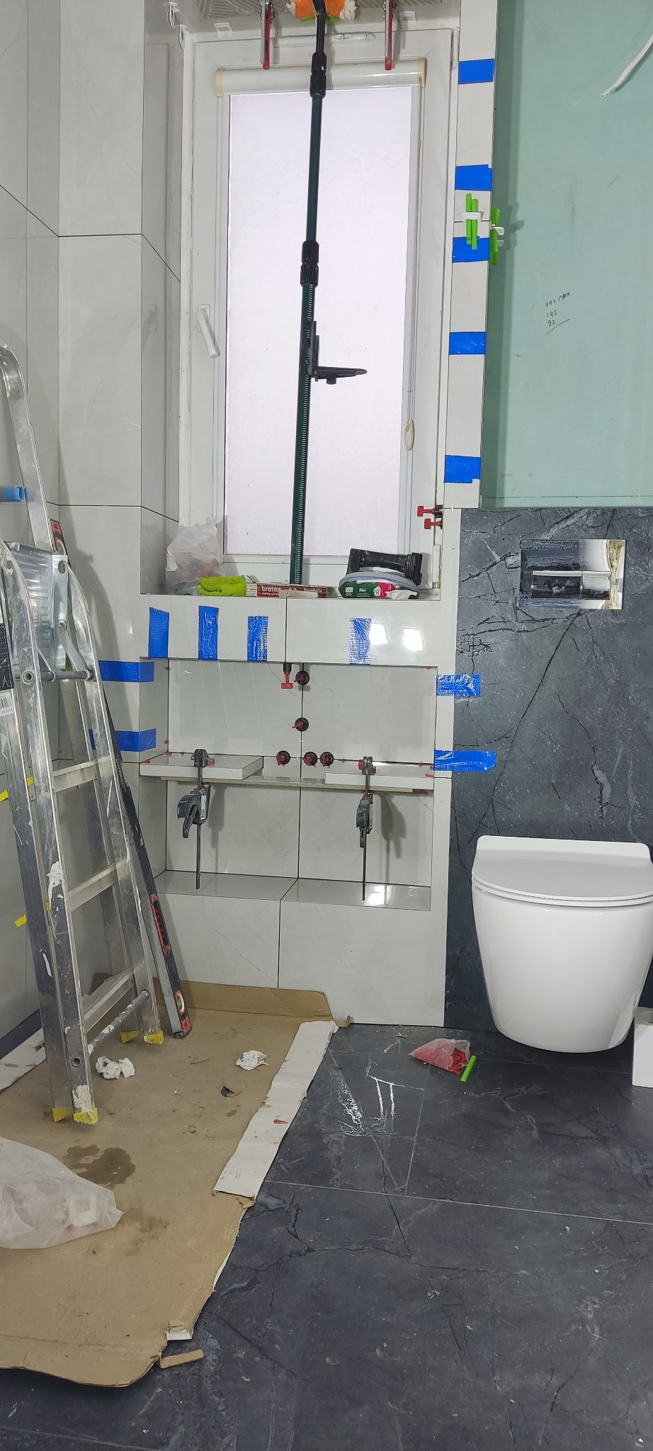 Łazienka w trakcie wykończenia: białe płytki na ścianie pod oknem, czarne płytki na podłodze, toaleta. Widoczne narzędzia glazurnicze i drabina.