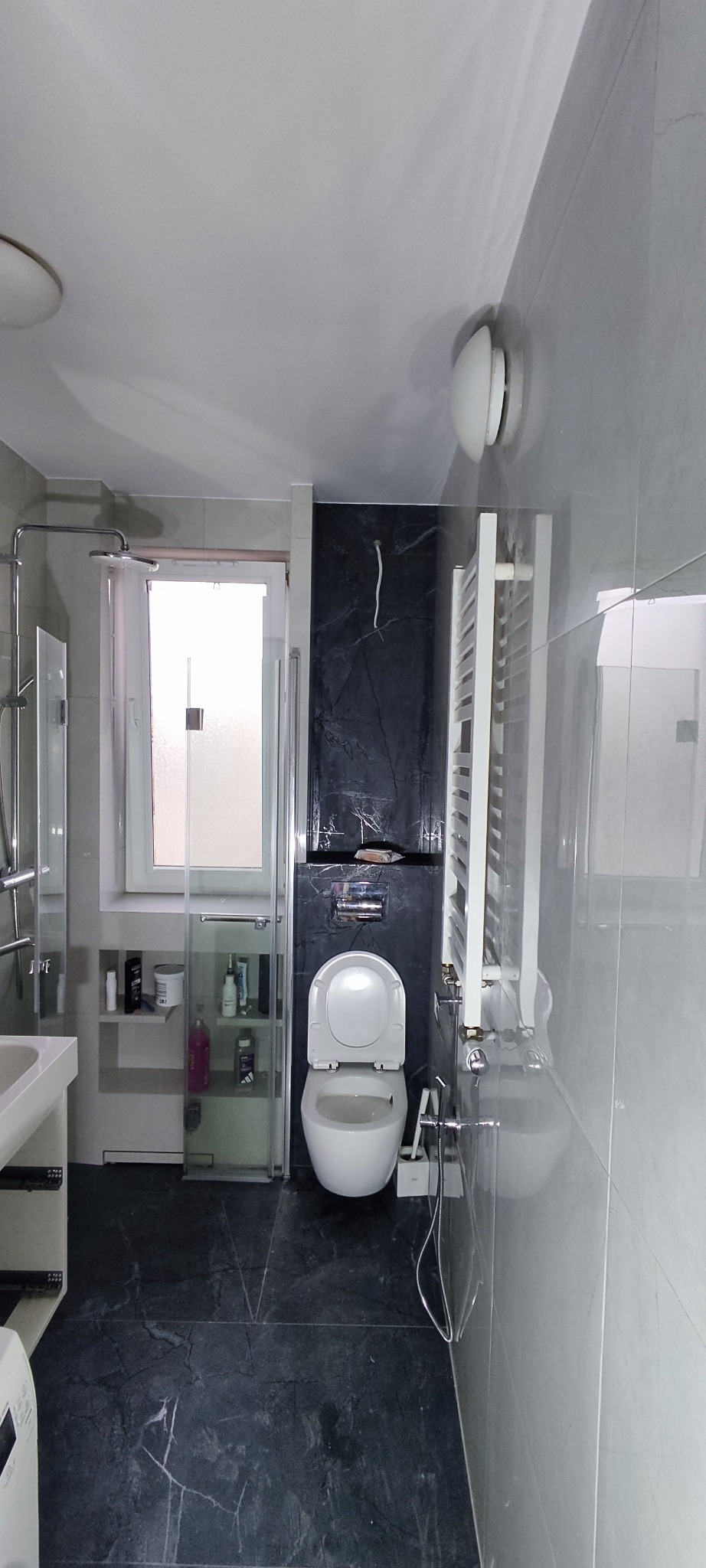 Nowoczesna łazienka z kabiną prysznicową, toaletą i grzejnikiem drabinkowym. Ciemna, marmurkowa podłoga i ściana za toaletą kontrastują z jasnymi płytkami.