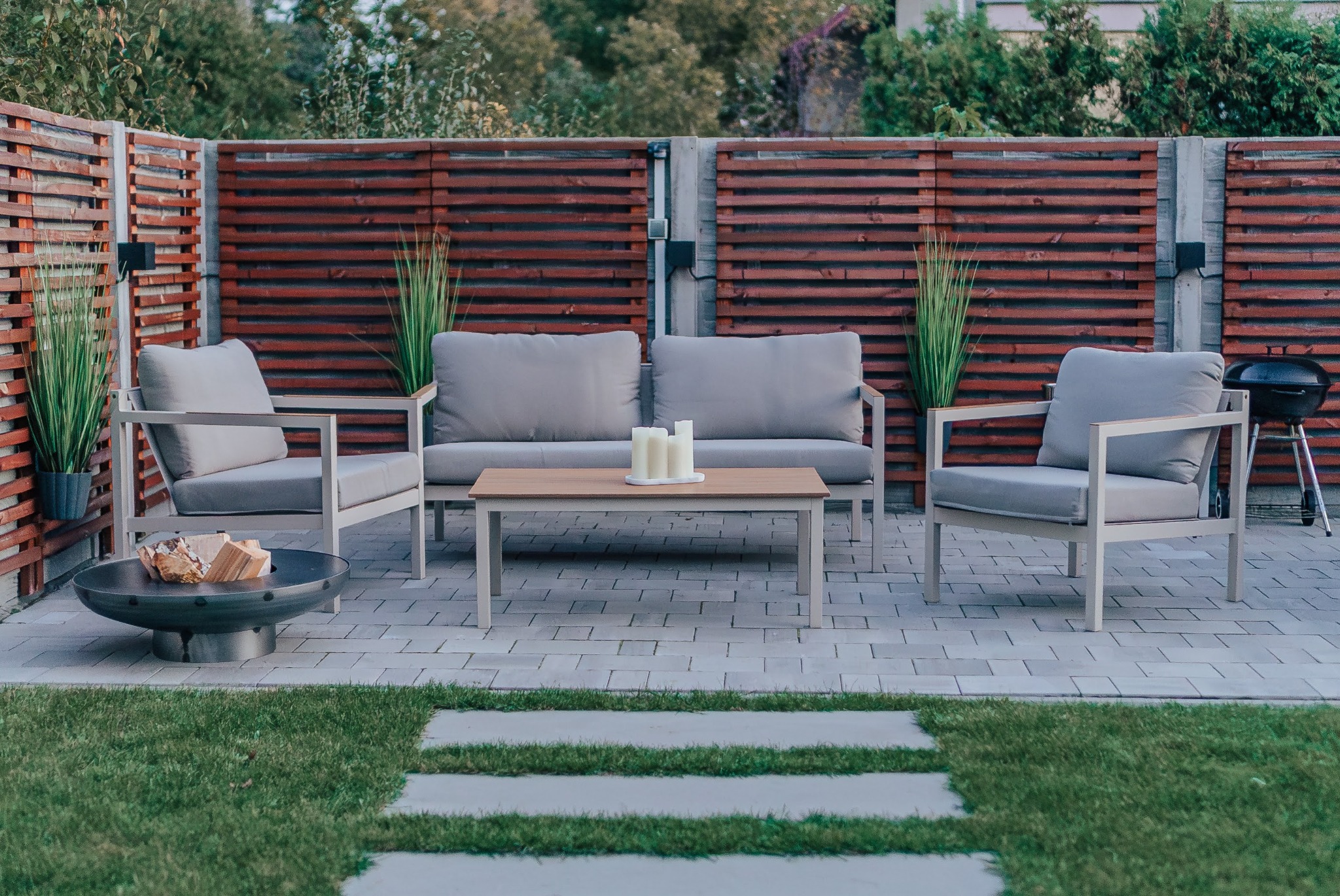 Patio z nowoczesnym zestawem mebli ogrodowych: sofa, fotele, stolik z trzema świecami. W tle drewniany płot i grill. Elementy dekoracyjne: trawa ozdobna, palenisko.