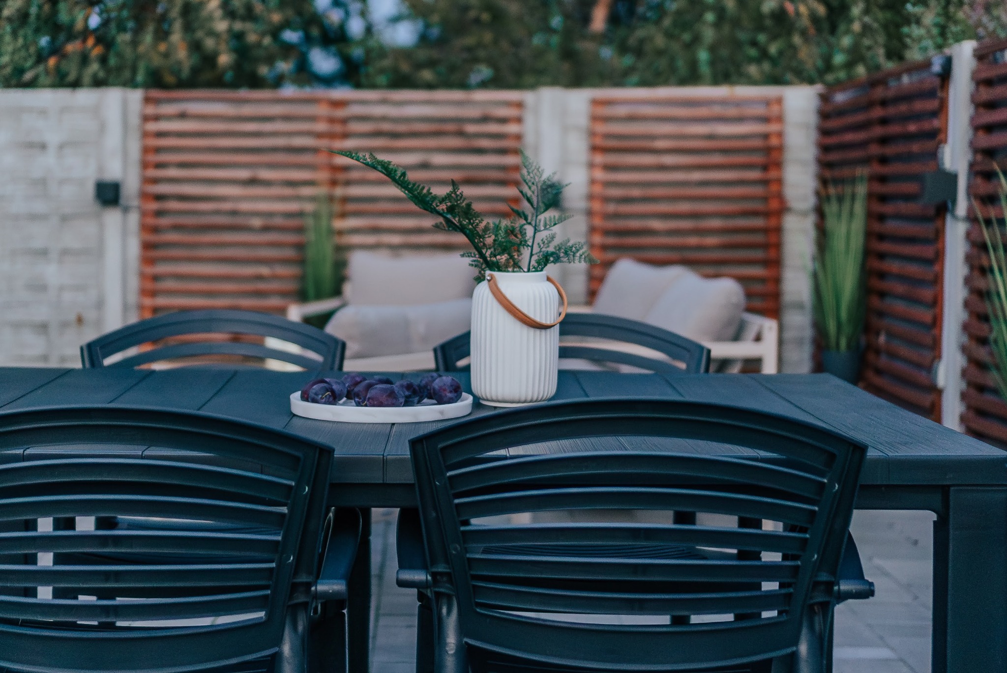 Stół ogrodowy z ciemnymi krzesłami, wazonem z paprotkami i talerzem śliwek na tle drewnianej pergoli i jasnych mebli wypoczynkowych.
