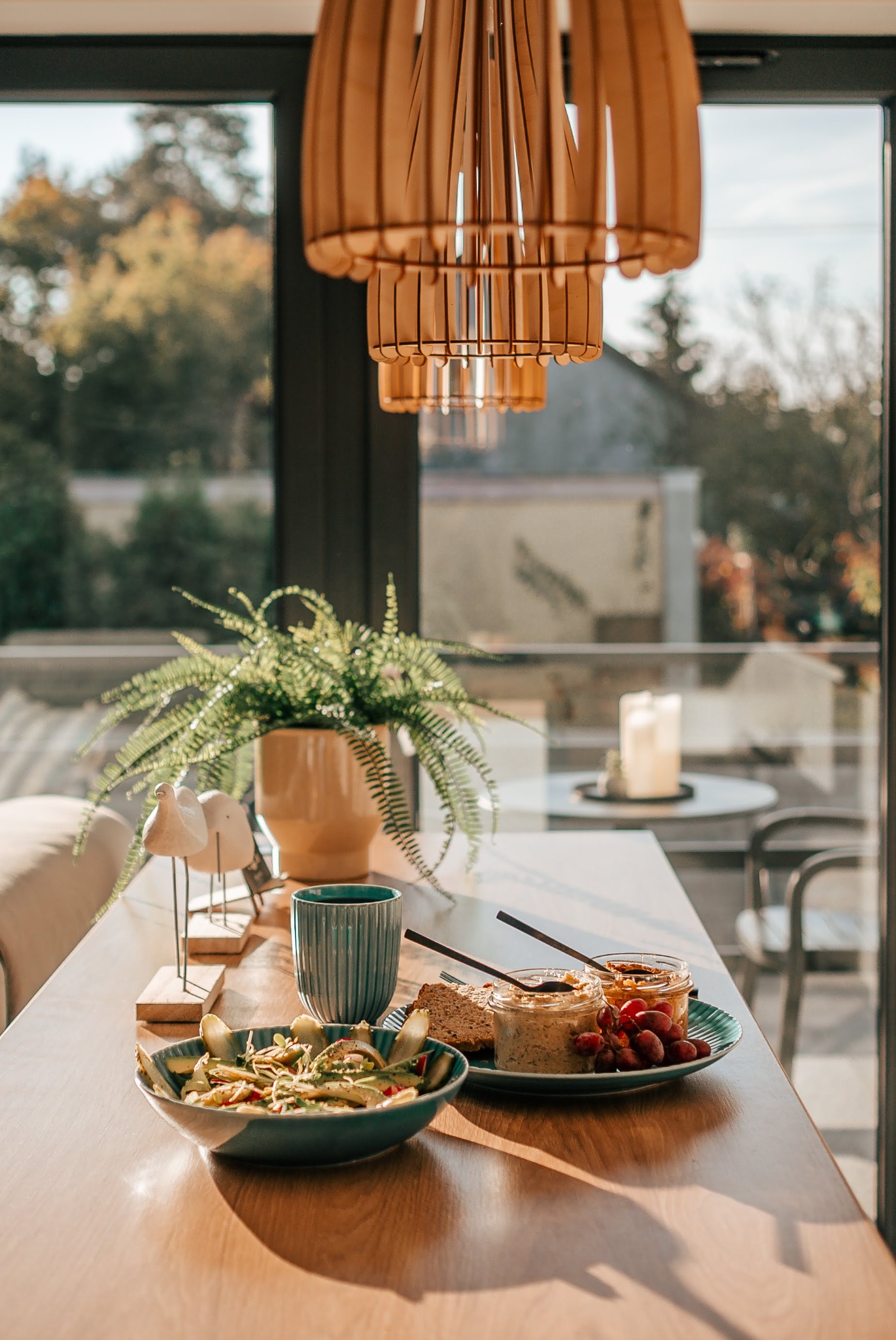 Stół z posiłkiem i dekoracjami w jasnym wnętrzu z dużym oknem, przez które widać zieleń. Nad stołem wisi nowoczesna, drewniana lampa.