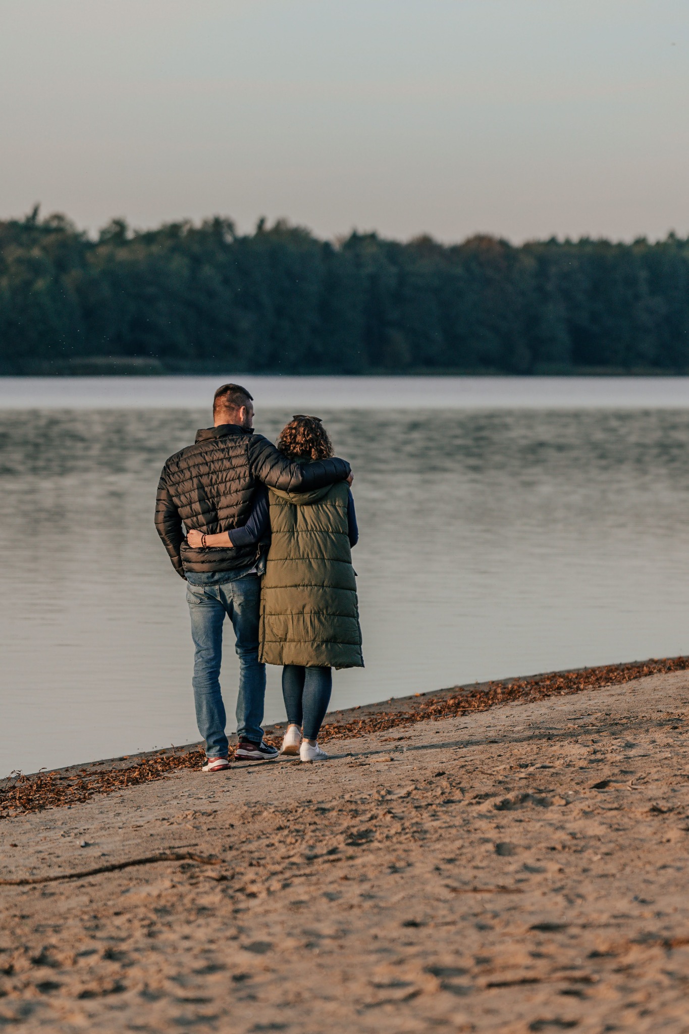 Para obejmuje się na brzegu jeziora, w tle drzewa i woda. Mężczyzna w czarnej kurtce, kobieta w długiej kamizelce. Spokojna atmosfera, widok z tyłu.