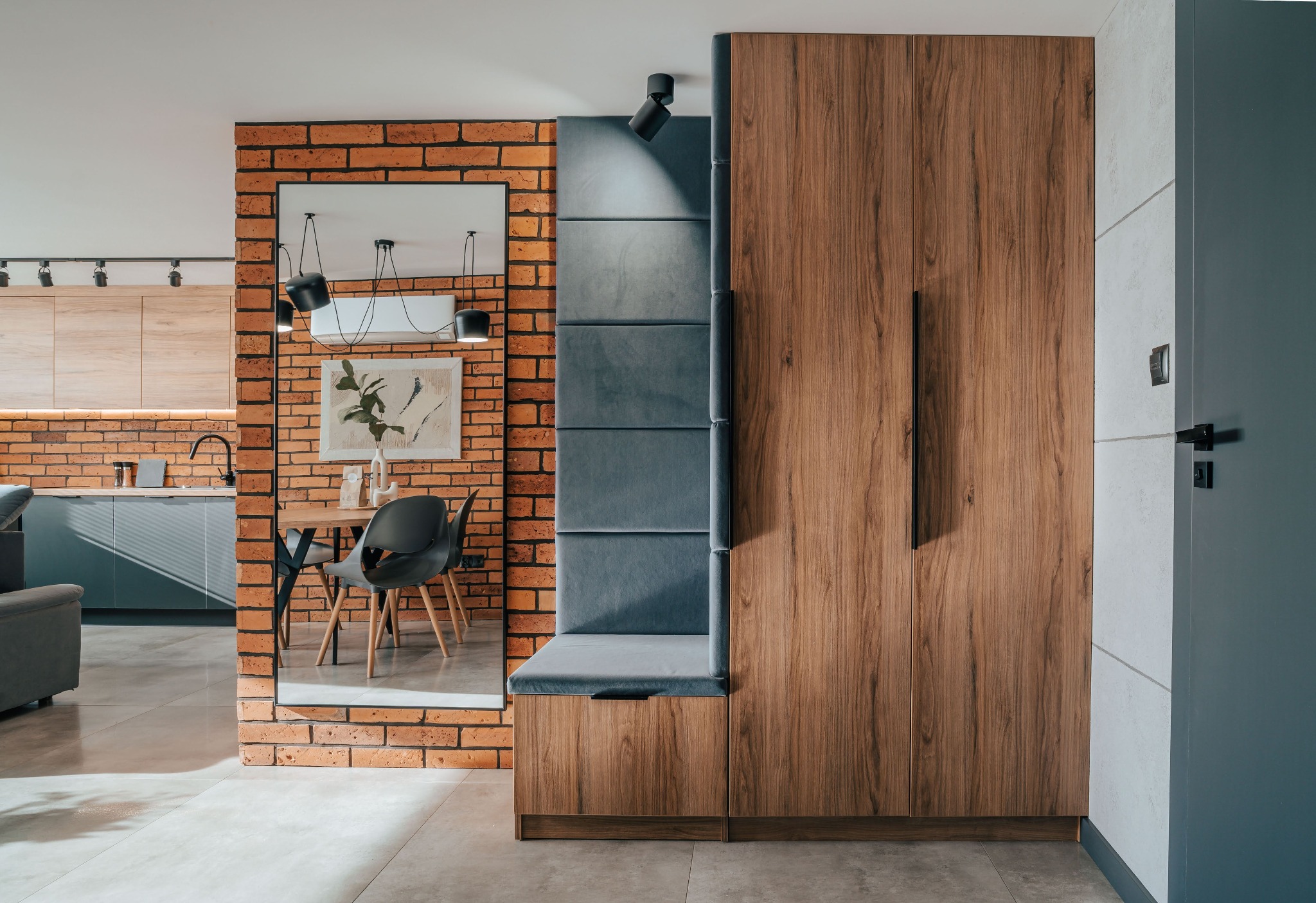 Nowoczesne wnętrze: zabudowa z drewnianymi frontami, szara tapicerka i ceglana ściana z lustrem odbijającym jadalnię. Minimalistyczny design, oświetlenie punktowe.