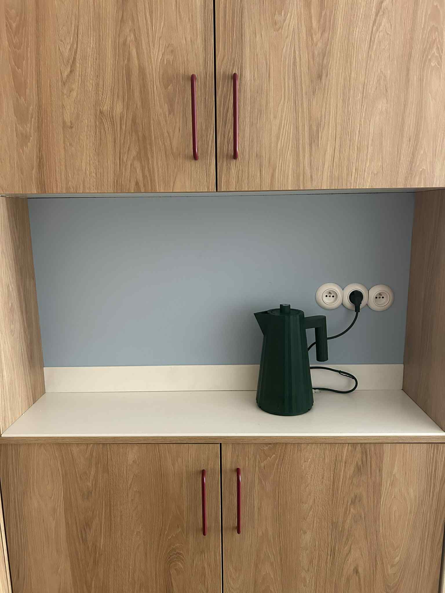 Wnęka kuchenna z drewnianymi szafkami, białym blatem i zielonym czajnikiem. Na ścianie trzy gniazdka elektryczne. Minimalistyczny design.