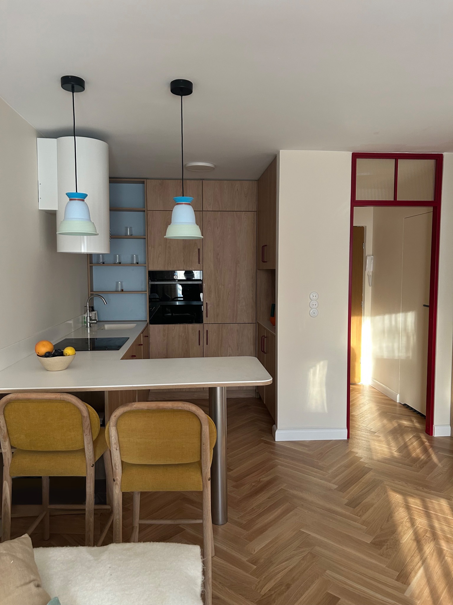 Jasna kuchnia z drewnianymi szafkami, blatem z owocami i żółtymi krzesłami barowymi. Parkiet w jodełkę i czerwona rama drzwi dodają koloru.