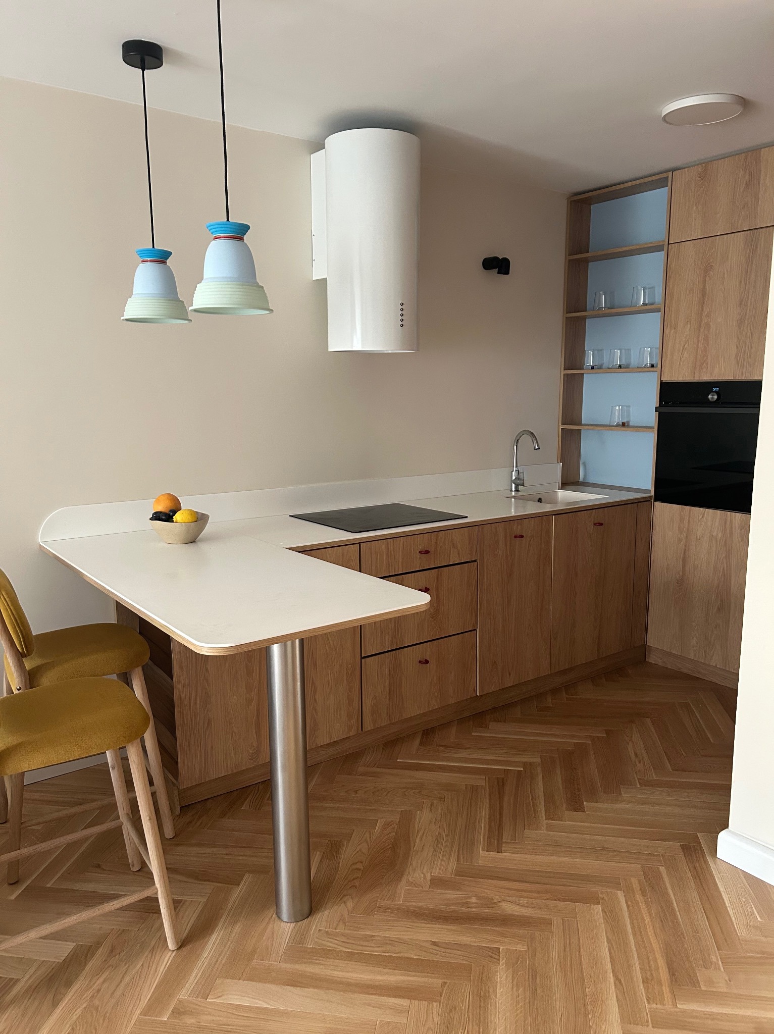 Nowoczesna kuchnia z drewnianymi szafkami, białym blatem i parkietem w jodełkę. Uwagę przykuwają designerskie lampy wiszące nad stołem.