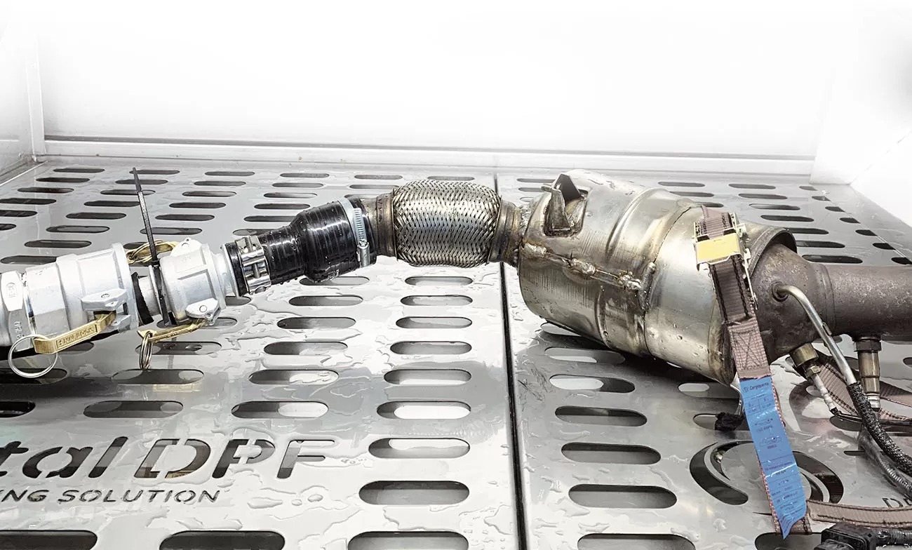 Filtr DPF podłączony do urządzenia czyszczącego, na metalowej perforowanej powierzchni. Widoczne złącza, przewody i elementy filtra. Detale czyszczenia DPF.