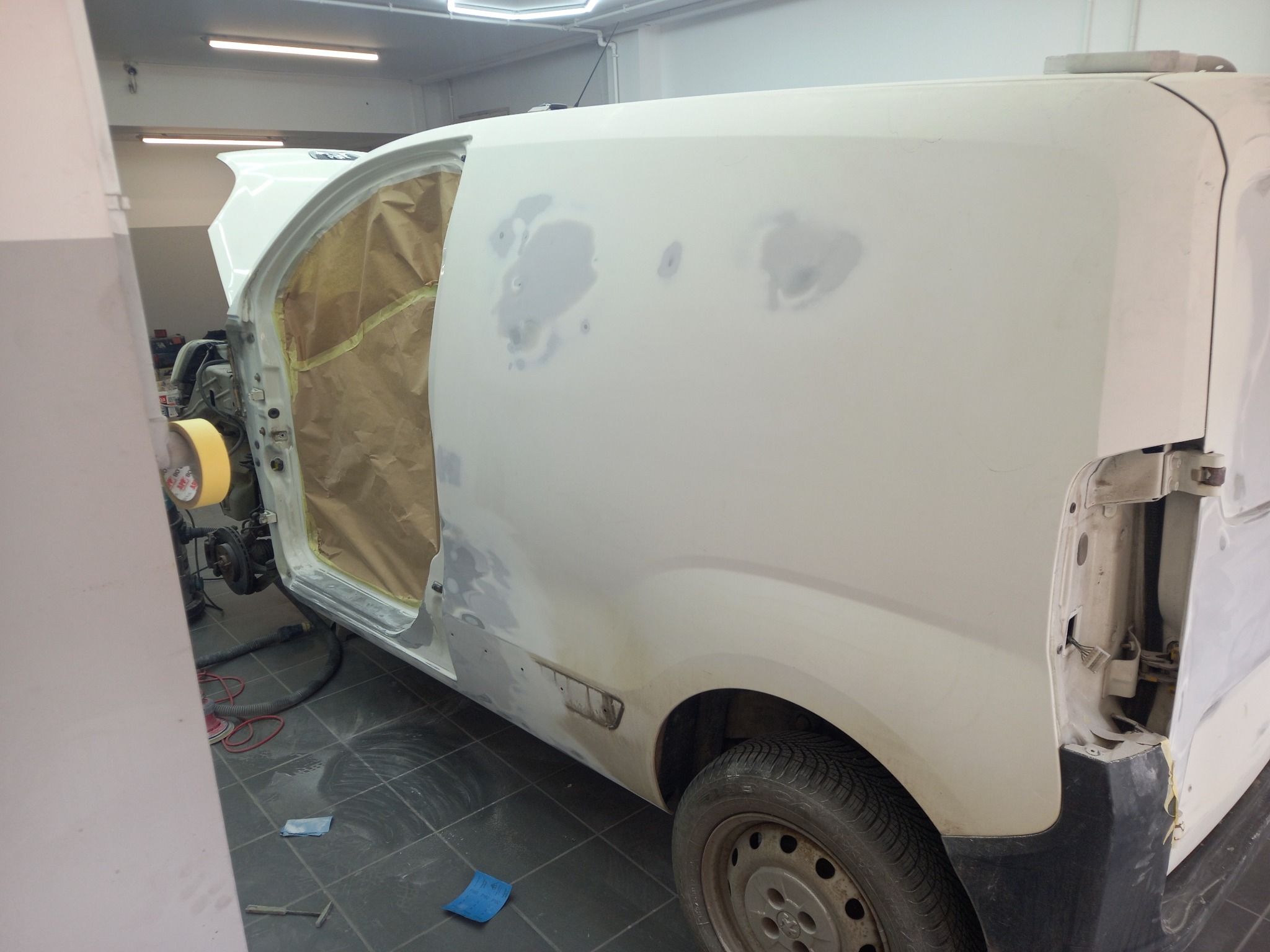 Biały van w warsztacie, przygotowany do lakierowania. Widoczne szpachlowane miejsca, otwarte drzwi zabezpieczone papierem i taśmą. Oświetlenie warsztatowe.