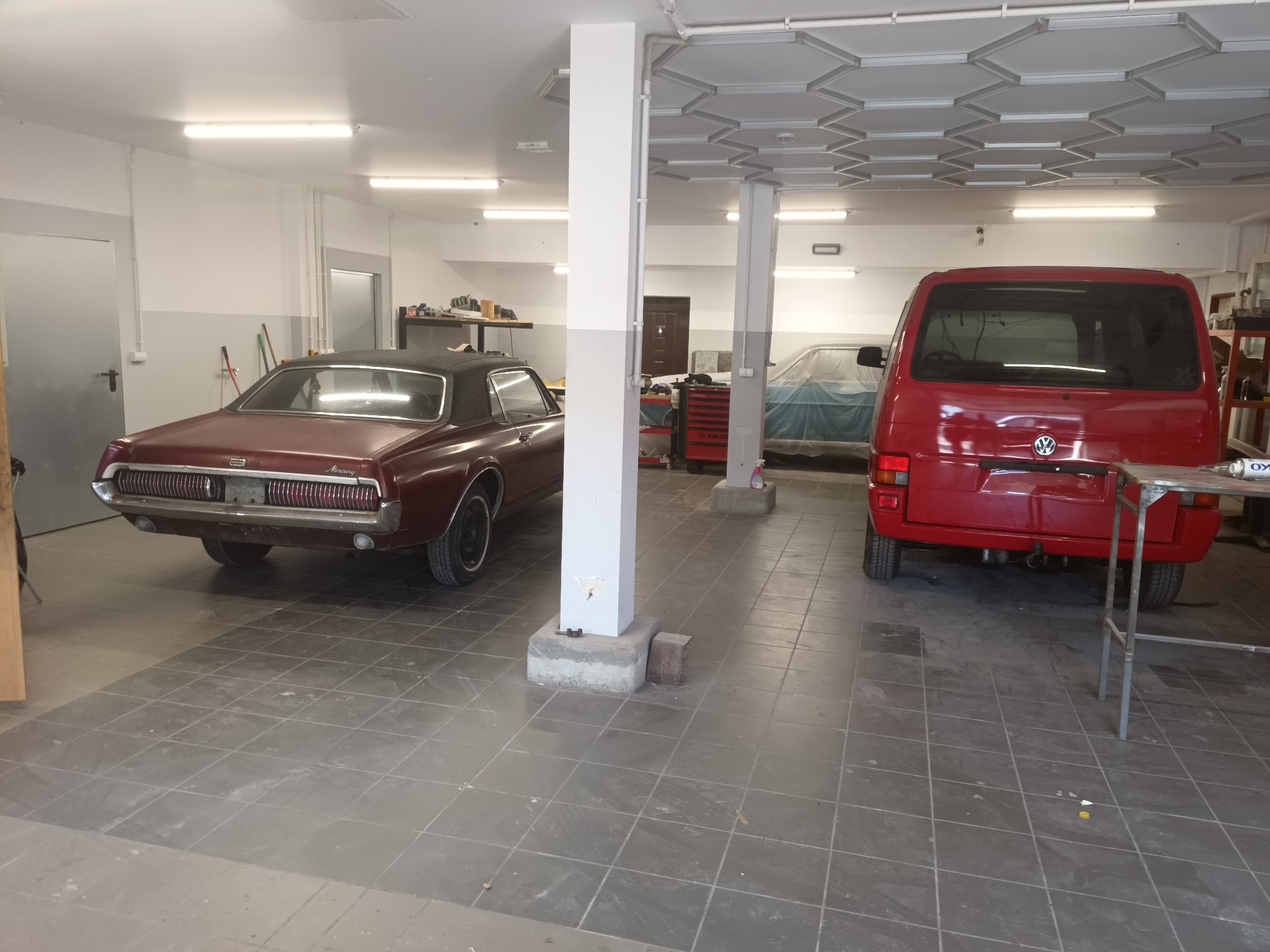 Wnętrze warsztatu samochodowego z bordo Mercury Cougar i czerwonym VW Transporter T4. W tle auto pod plandeką, narzędzia, płytki na podłodze i kolumny podtrzymujące strop.