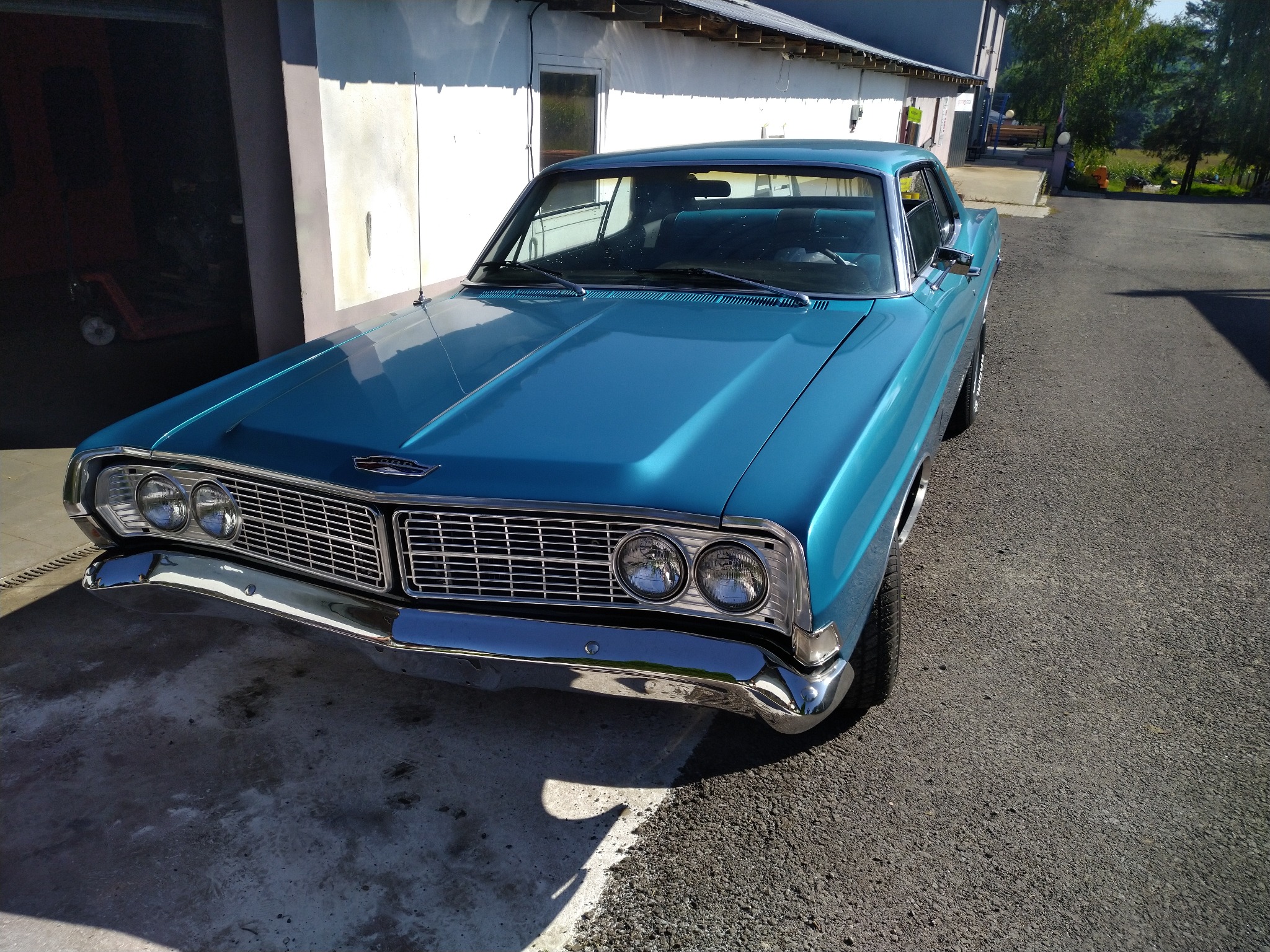Błękitny, odrestaurowany Ford Galaxie z chromowanym grillem i reflektorami, zaparkowany na asfalcie przed garażem. Widoczne odbicia światła na karoserii.
