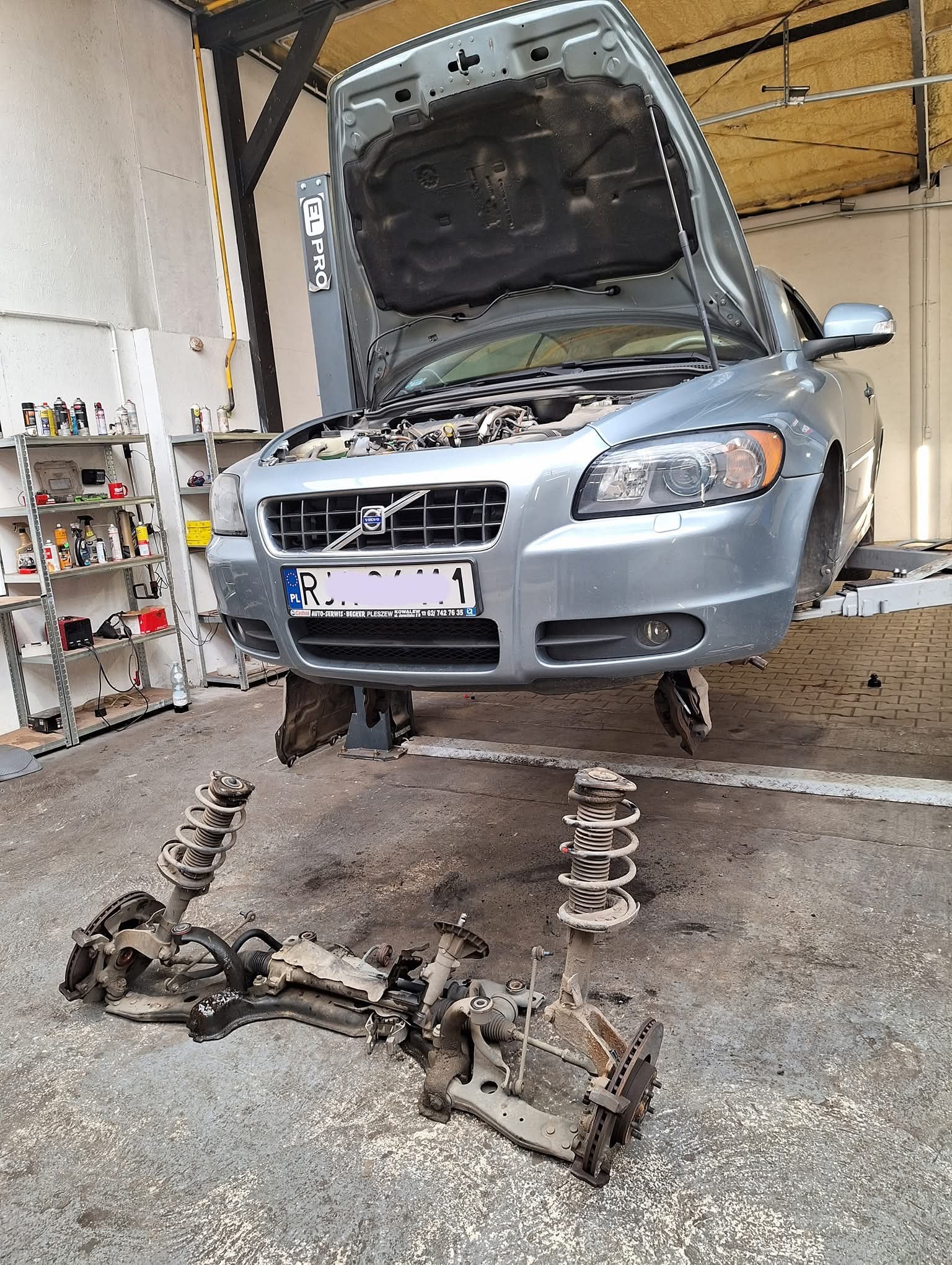Szary samochód Volvo na podnośniku w warsztacie z otwartą maską. Zdemontowane elementy zawieszenia leżą na podłodze. Widoczne regały z narzędziami.
