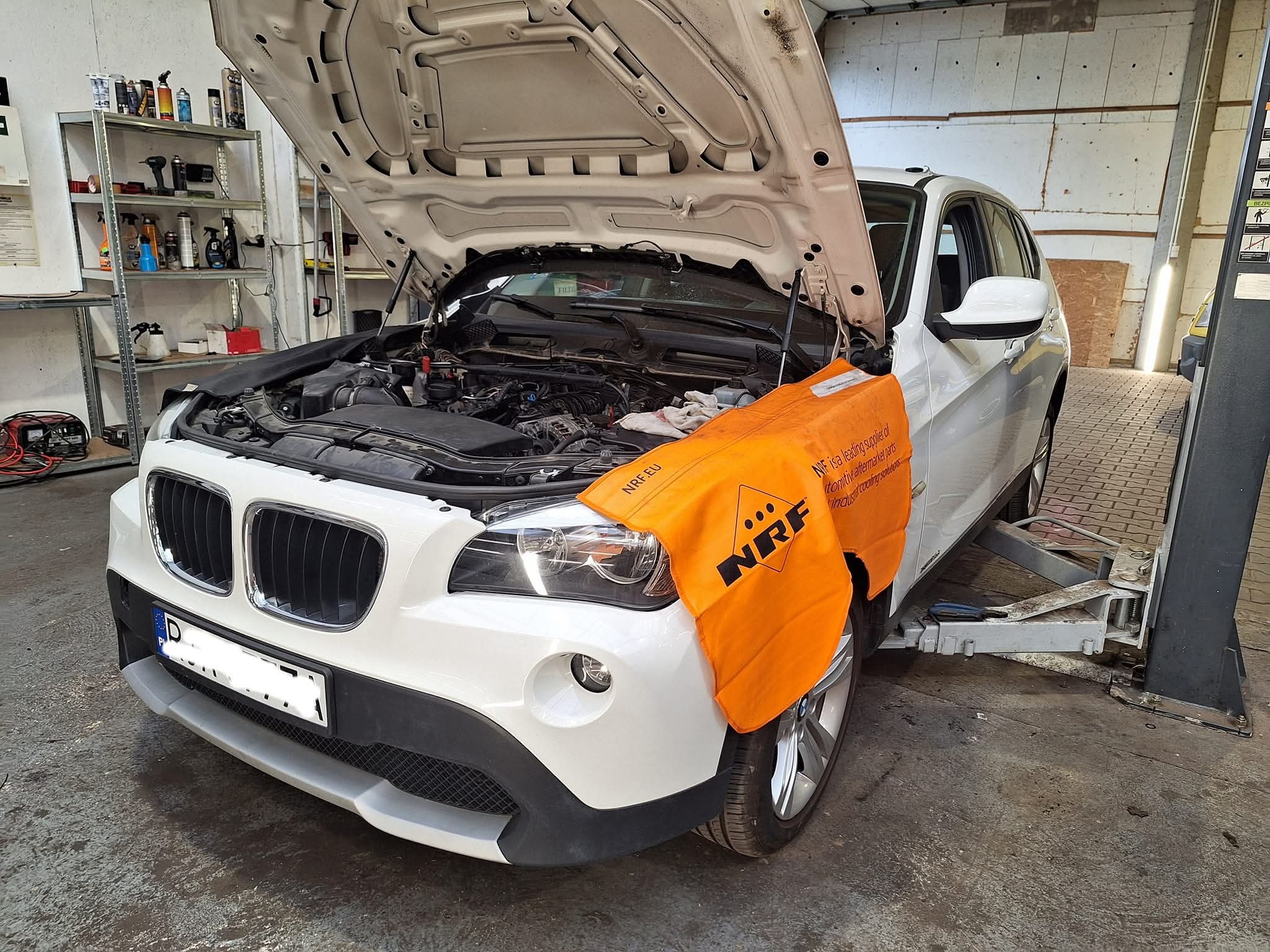 Białe BMW X1 z otwartą maską na podnośniku w warsztacie. Pomarańczowa osłona NRF na błotniku. Widoczny silnik i narzędzia na półkach w tle.