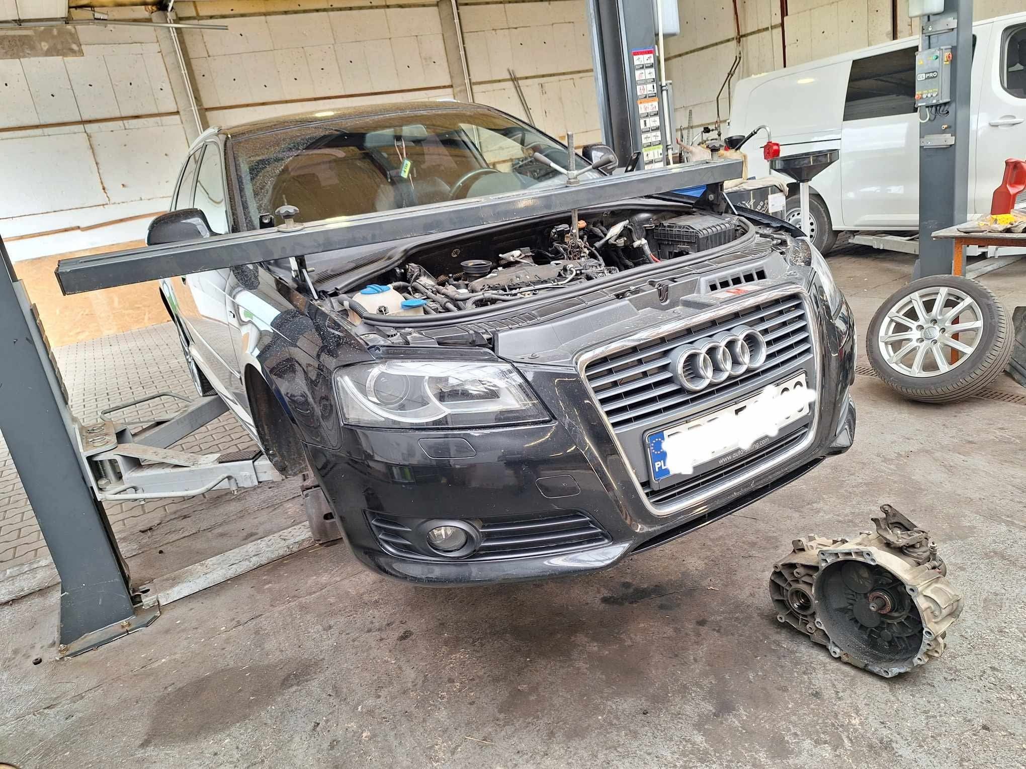 Czarny Audi A3 na podnośniku warsztatowym z otwartą maską i zdemontowaną skrzynią biegów obok, w tle widoczny bus i opona.