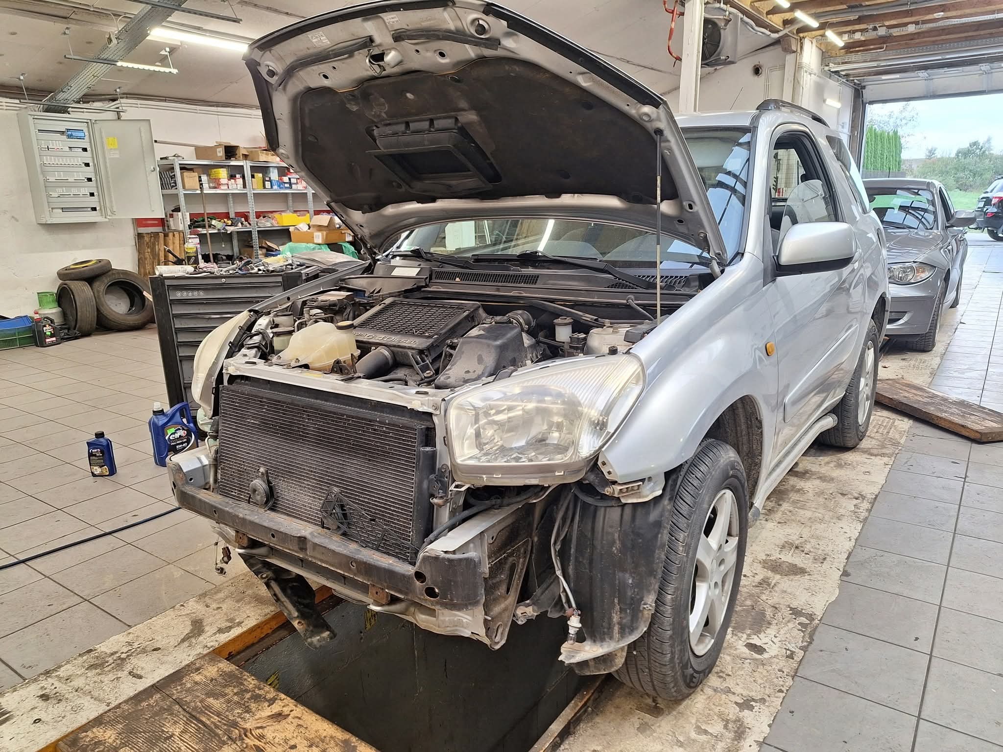 Srebrny SUV z otwartą maską w warsztacie, widoczny silnik i chłodnica. Brak przedniego zderzaka. W tle narzędzia i półki z częściami. Podnośnik warsztatowy.
