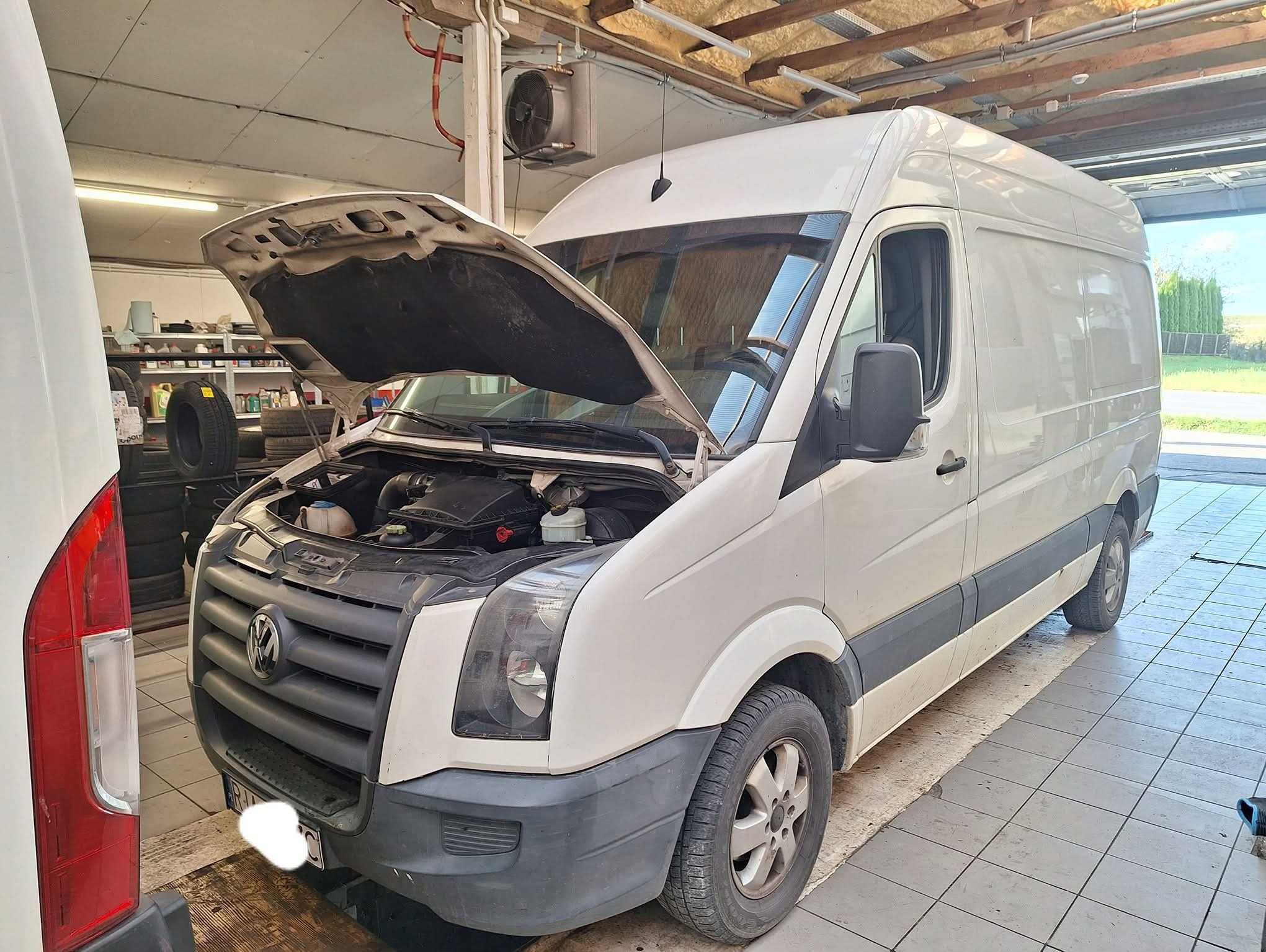 Biały van Volkswagen Crafter z otwartą maską w warsztacie samochodowym, widoczne opony w tle. Diagnostyka lub naprawa silnika w toku.