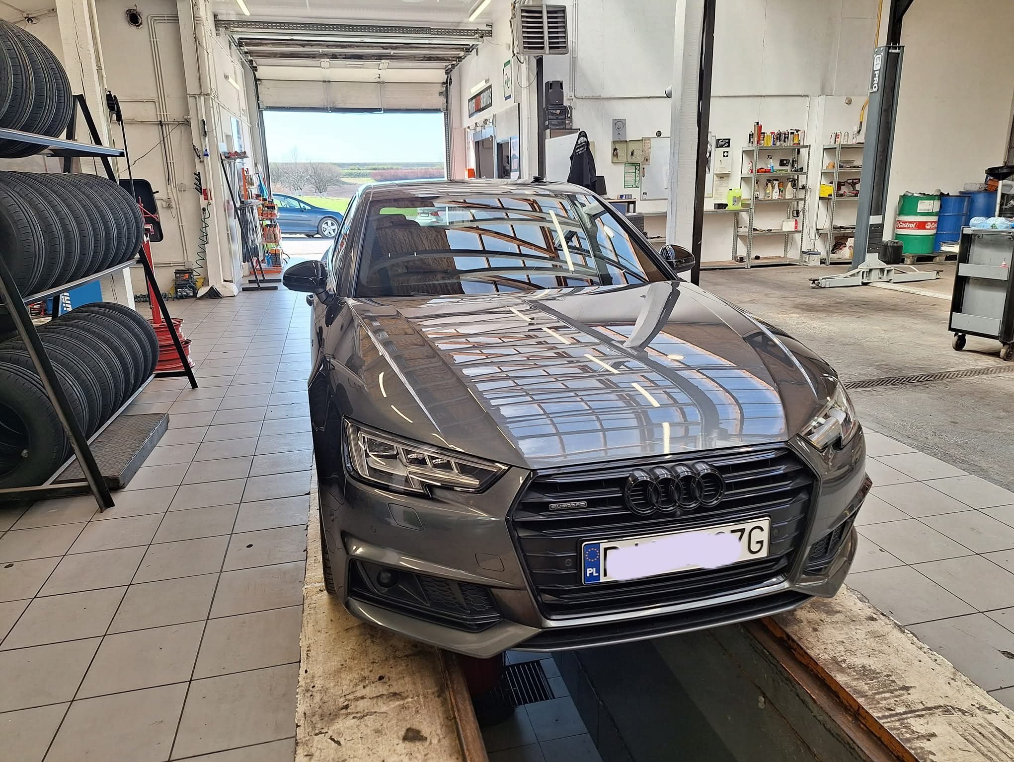 Szary samochód Audi A4 na podnośniku w warsztacie samochodowym, widoczne opony na regale i otwarte wrota garażu z widokiem na zewnątrz.