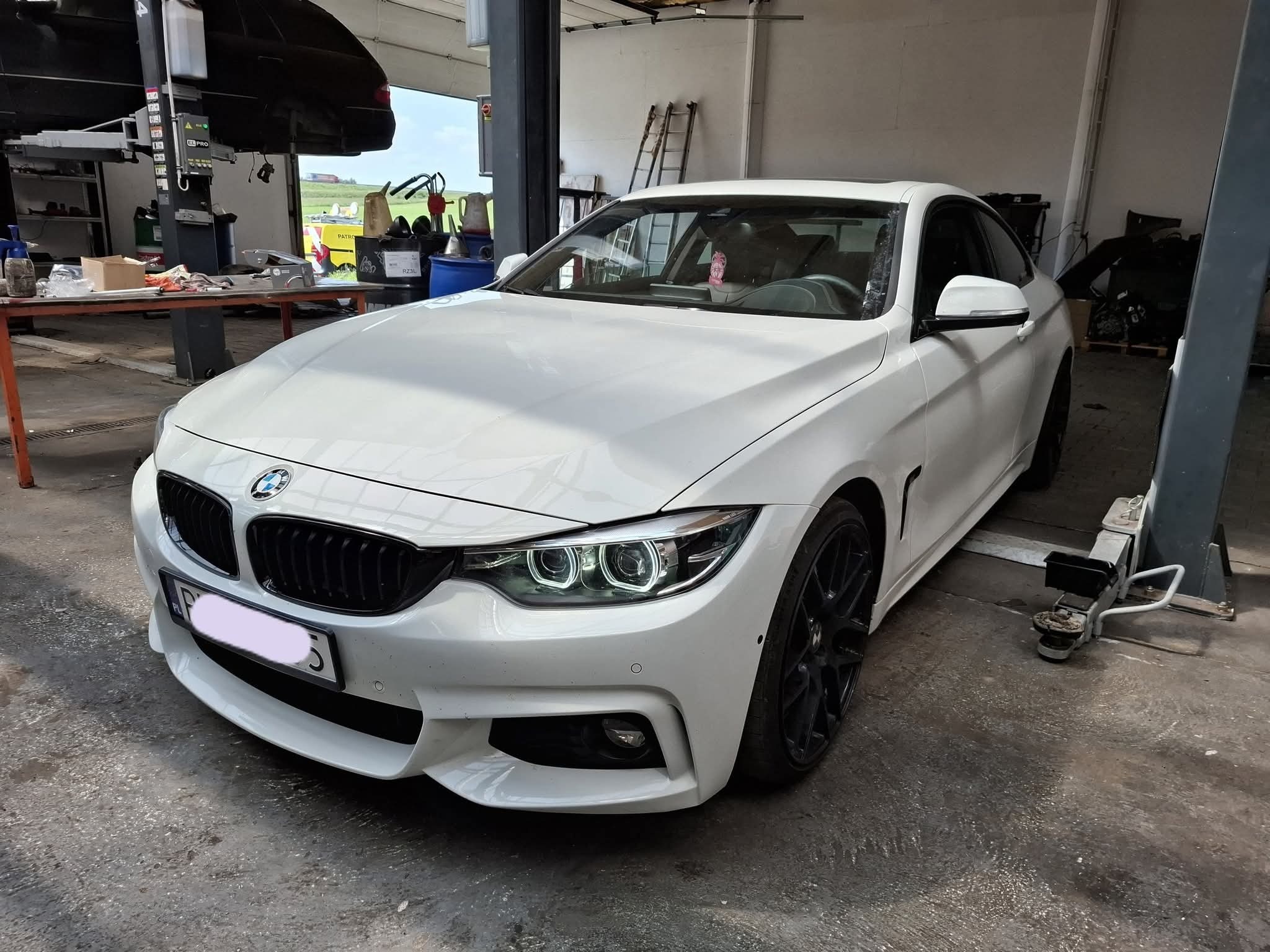 Białe BMW serii 4 Coupe na podnośniku w warsztacie, widok z przodu, czarne felgi, światła LED, w tle podnośnik z innym autem i narzędzia.