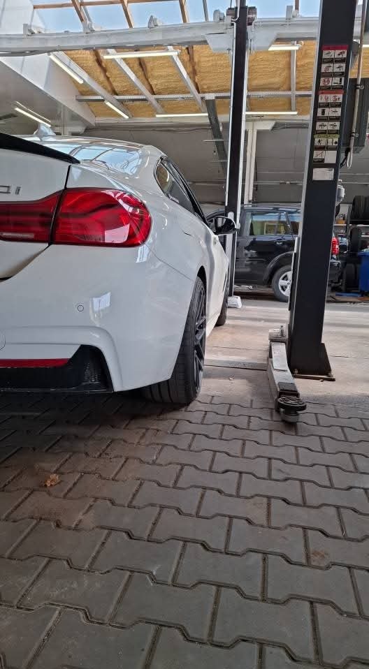 Białe auto na podnośniku w warsztacie, widok z tyłu, z bliska. Widoczne oświetlenie sufitowe i podłoga z kostki brukowej.