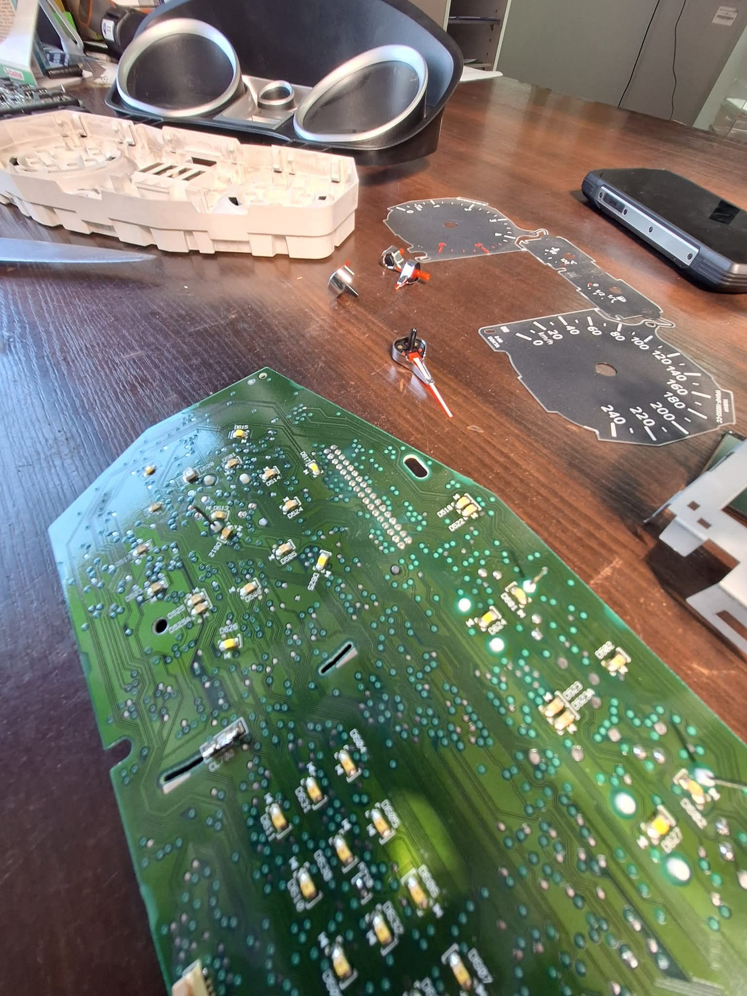 Rozłożony panel zegarów samochodowych z widoczną płytką drukowaną, tarczami prędkościomierza i obrotomierza, wskazówkami oraz elementami obudowy na drewnianym stole.