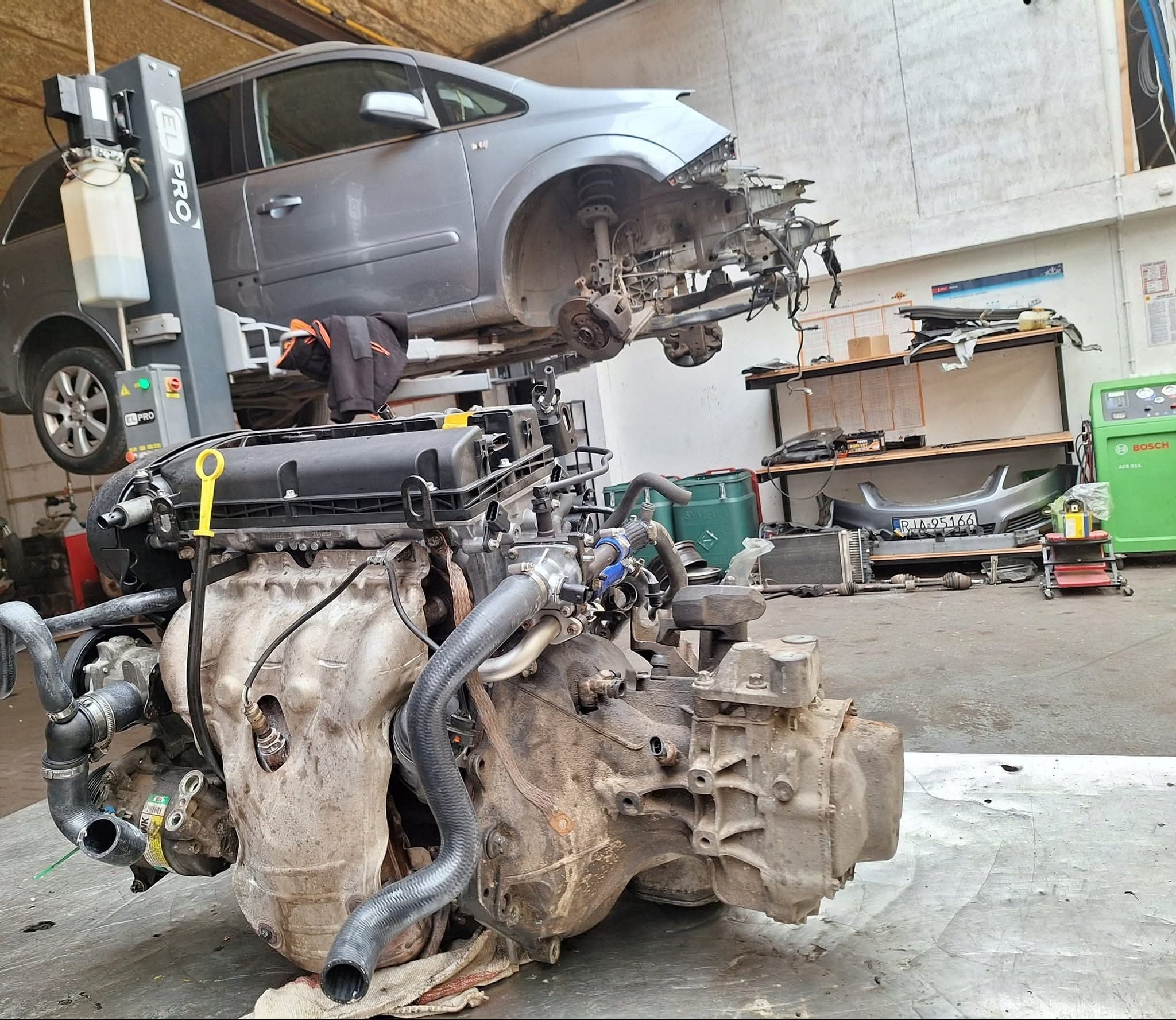 Silnik samochodowy z osprzętem na stole warsztatowym, w tle podnośnik z rozmontowanym autem. Widoczne narzędzia i części. Warsztat mechaniczny w trakcie pracy.