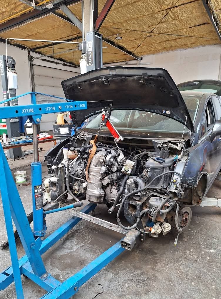 Rozłożony silnik samochodu zawieszony na podnośniku w warsztacie. Widoczne elementy karoserii, wnętrze silnika i narzędzia warsztatowe. Oświetlenie warsztatowe.