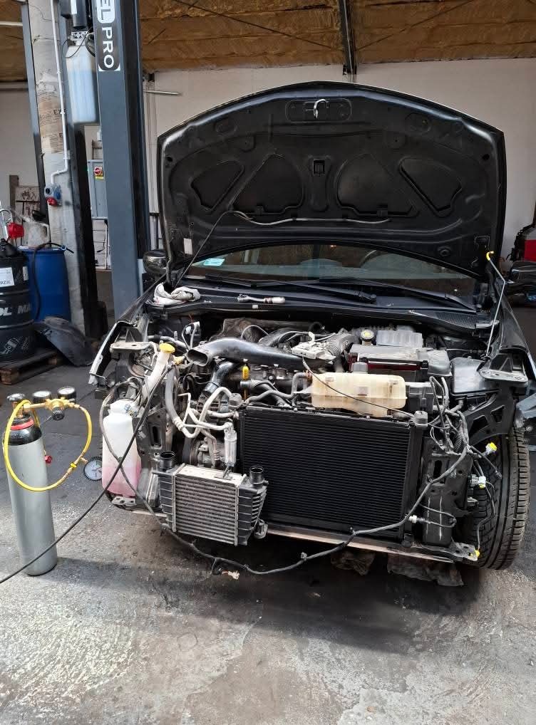 Czarny samochód z otwartą maską w warsztacie, widoczny silnik i chłodnica. Podłączony sprzęt diagnostyczny, butla z gazem. Auto na podnośniku.