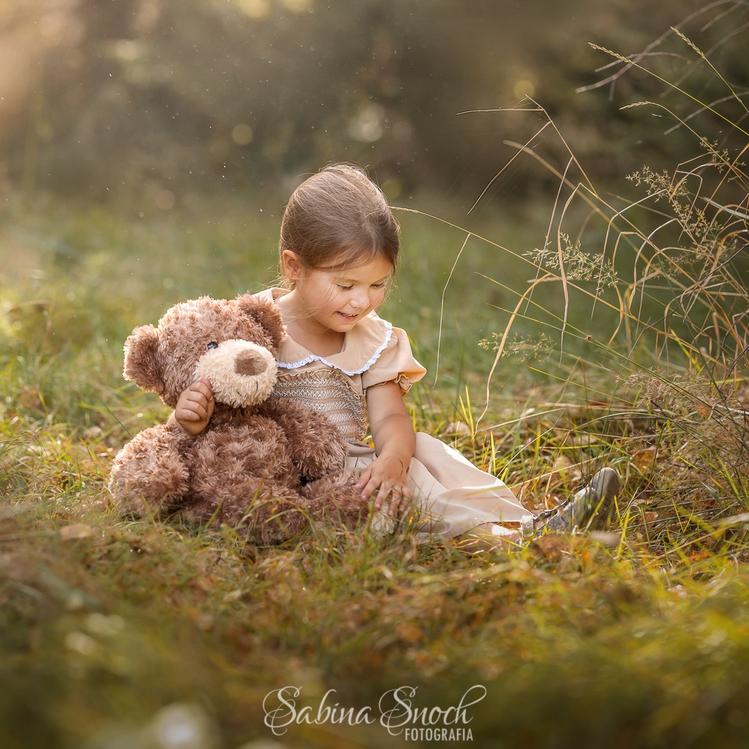 Dziewczynka w beżowej sukience siedzi na trawie, przytulając pluszowego misia w promieniach słońca, z widocznym logo fotografa w dolnej części kadru.