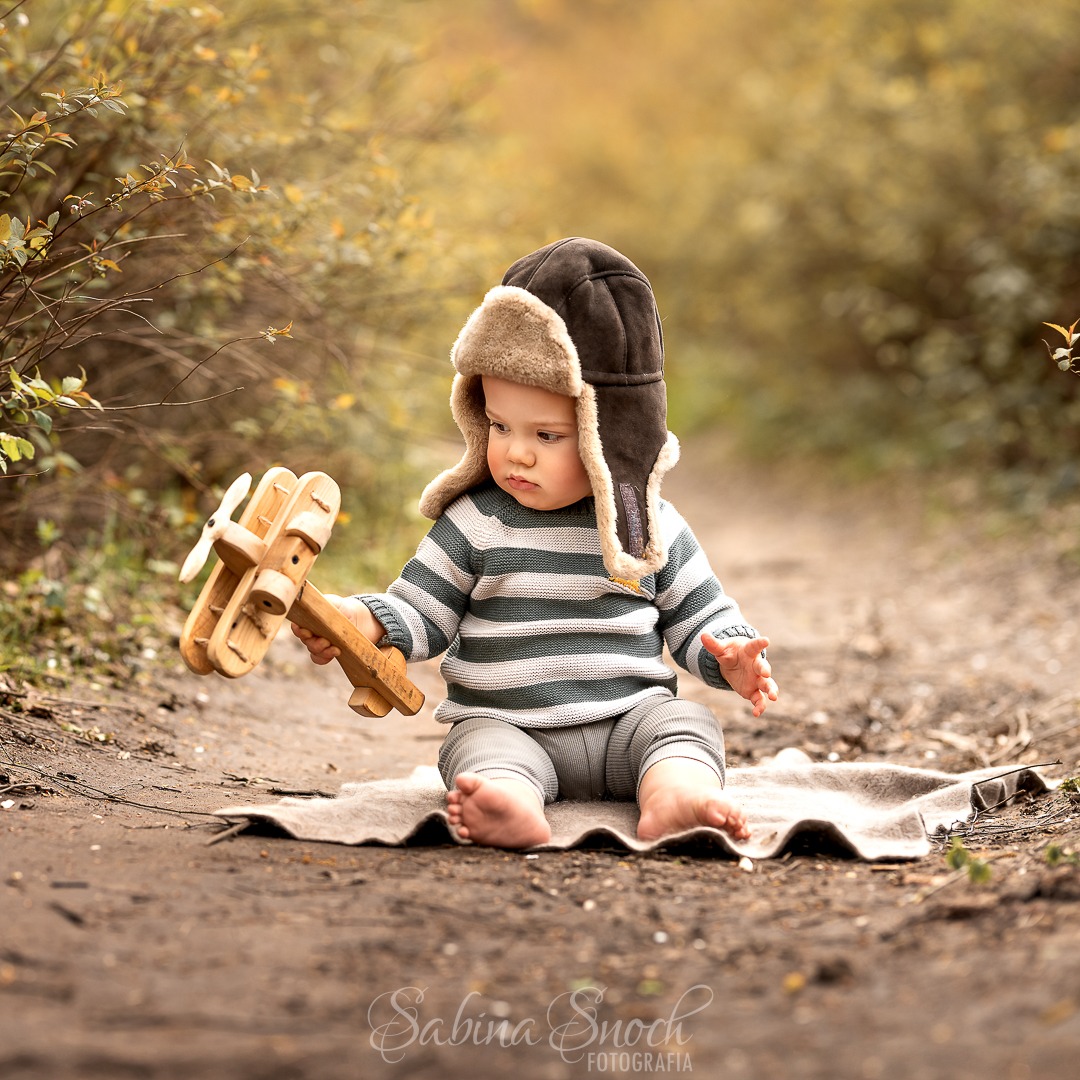 Uroczy niemowlak w czapce pilota, siedzący na kocu w plenerze, trzyma drewniany samolot. Jesienna sceneria i miękkie światło tworzą ciepłą atmosferę.