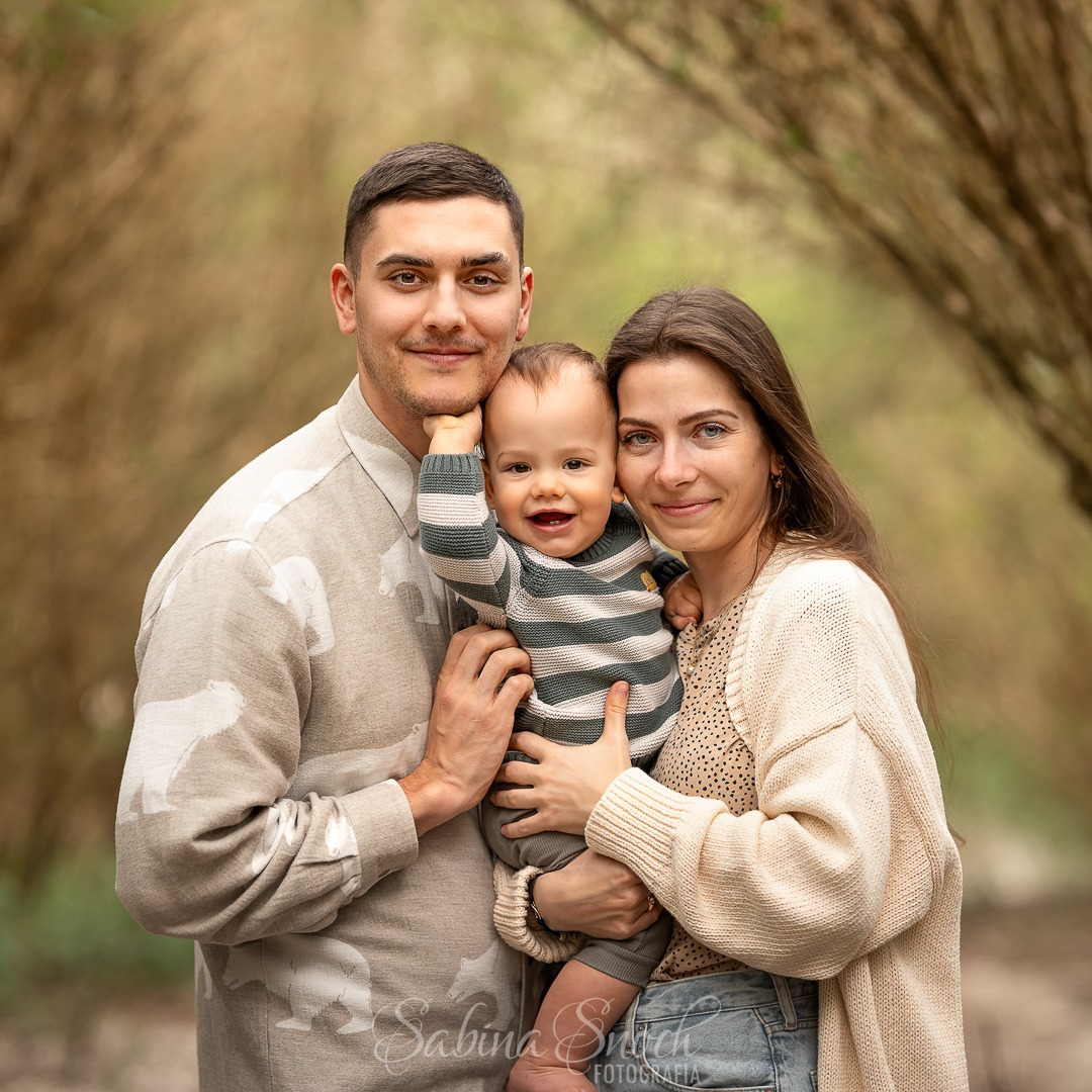 Uśmiechnięta rodzina: tata w koszuli w niedźwiedzie, mama w kardiganie i syn w paski, pozują na tle alei drzew w ciepłym świetle, tworząc portret rodzinny.