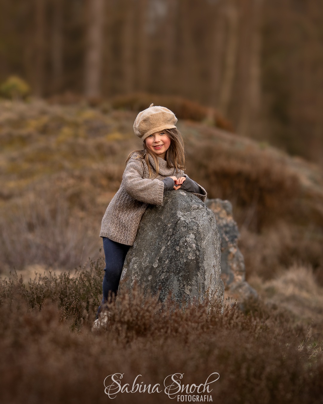 Uśmiechnięta dziewczynka w beretce, oparta o kamień na tle brązowej roślinności i rozmytego lasu. Naturalna sesja w plenerze.