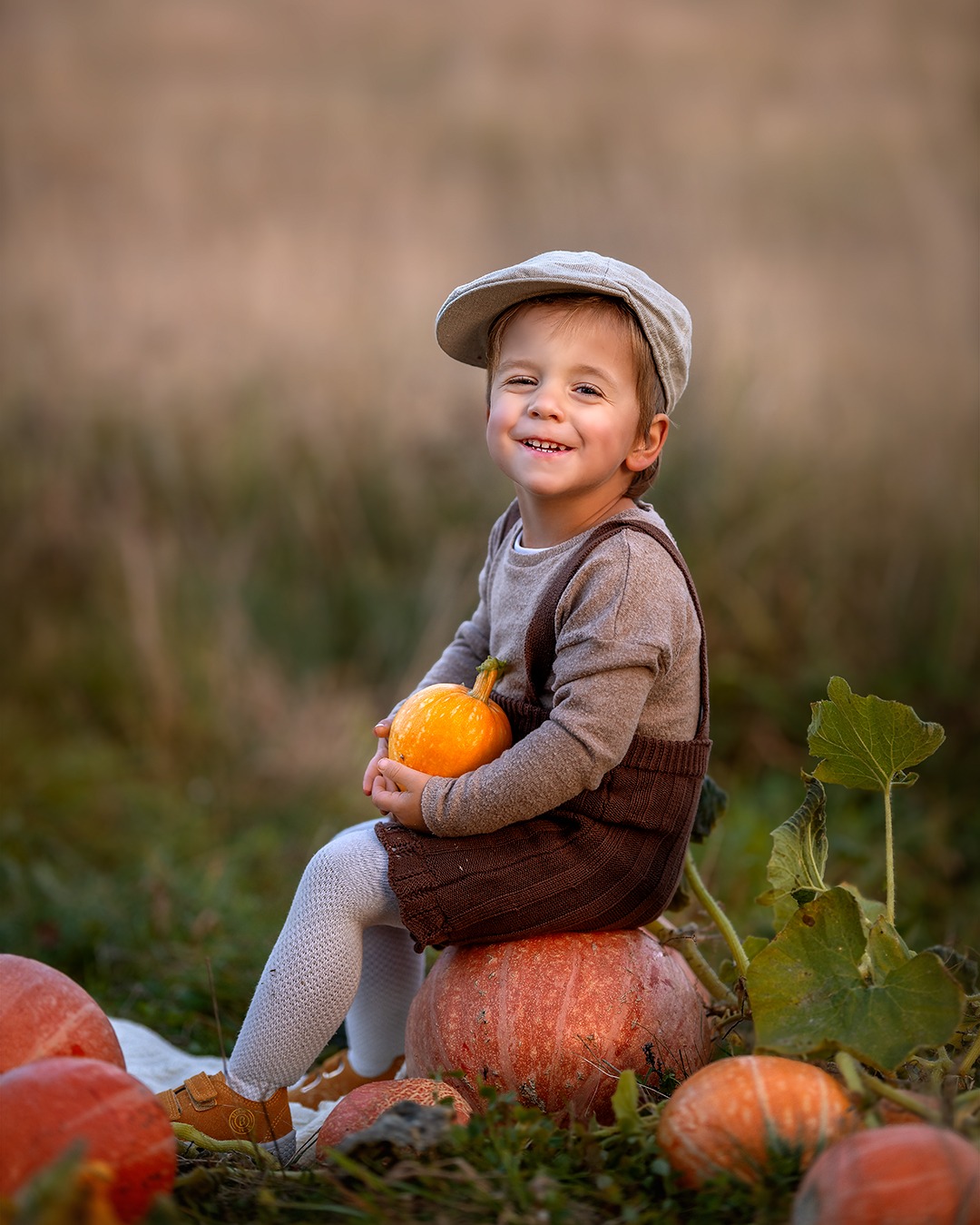 Uśmiechnięty chłopiec w kaszkiecie siedzi na dyni, trzymając małą dynię. Jesienna sceneria, miękkie światło, naturalne tło. Urocza sesja w plenerze.