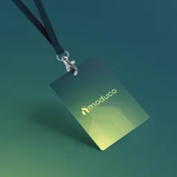 Identyfikator z logo 'moduco' zawieszony na smyczy, z gradientem od zieleni do żółci, leżący na zielonym tle. Metalowy karabińczyk i teksturowana smycz.