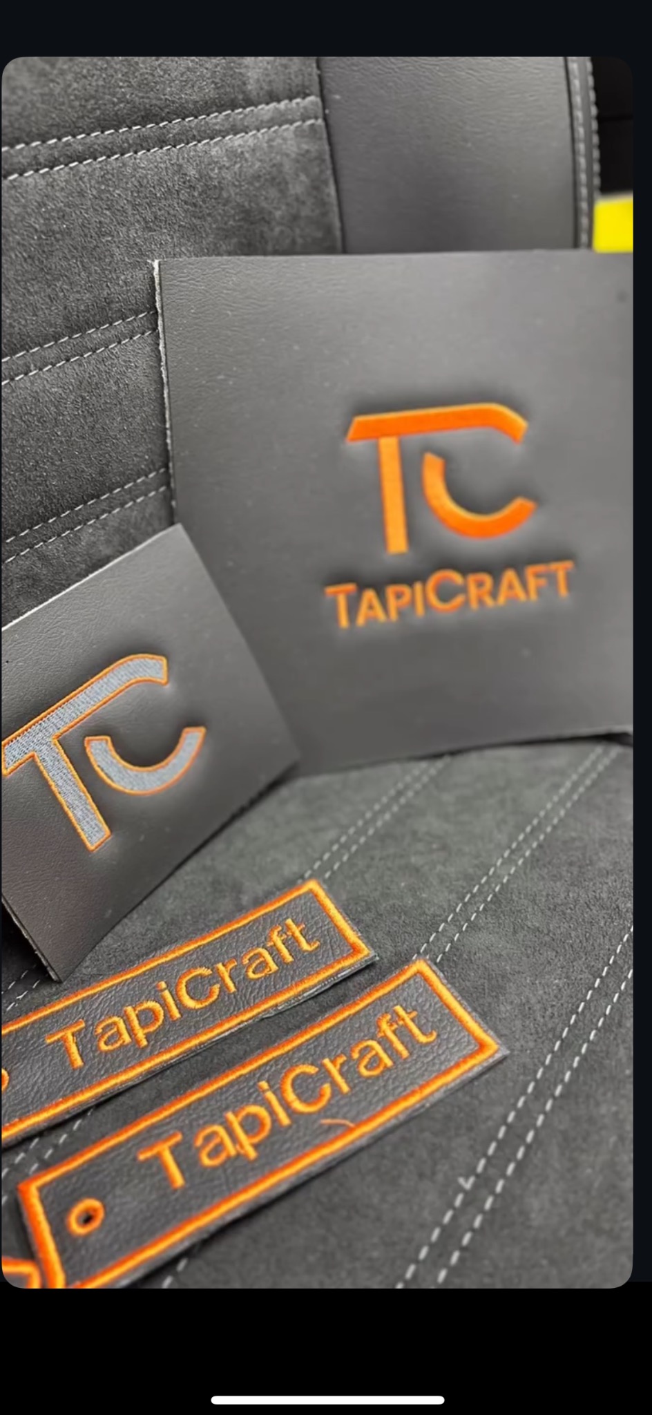 Szare skórzane próbki z pomarańczowym logo TapiCraft na tle tapicerki z szarymi przeszyciami.