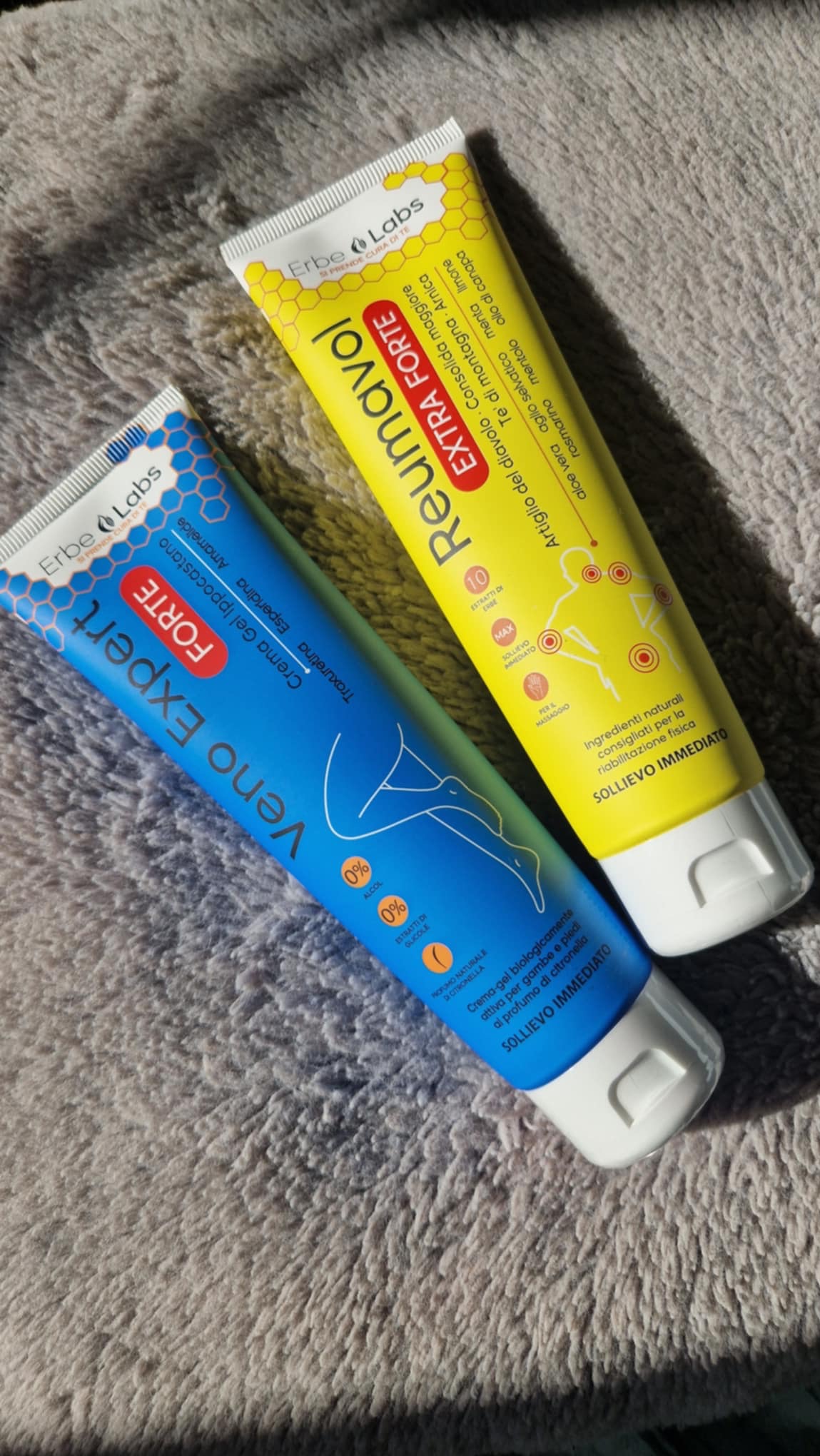 Dwie tubki kremów Erbe Labs: niebieska Veno Expert Forte z grafiką nóg i żółta Reumavol Extra Forte z listą składników. Tło to teksturowana, szara powierzchnia.