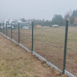 Ogrodzenia i Zieleń sp. z o.o. - Długi, szary panel ogrodzeniowy z podmurówką betonową, ciągnący się wzdłuż pola w mglisty dzień. W tle zabudowania i drzewa.