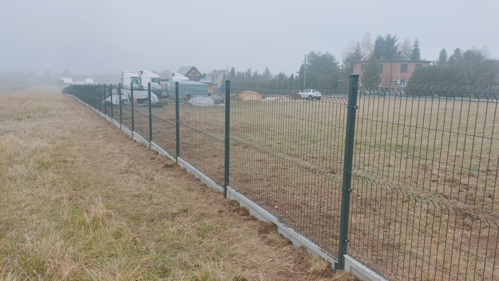 Długi, szary panel ogrodzeniowy z podmurówką betonową, ciągnący się wzdłuż pola w mglisty dzień. W tle zabudowania i drzewa.