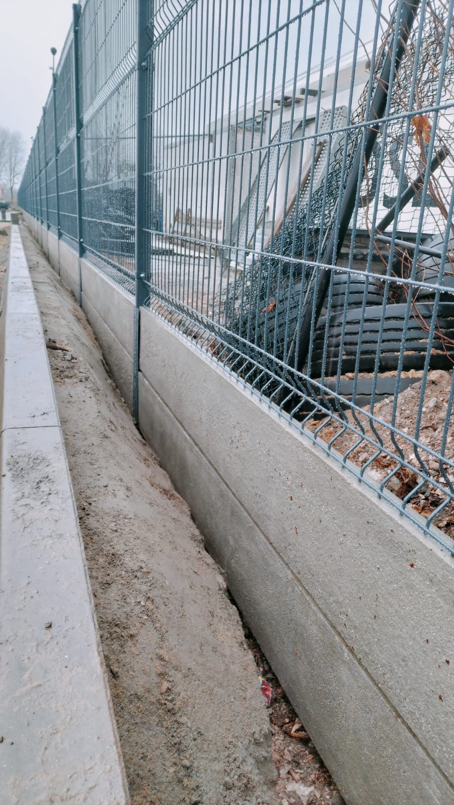 Nowoczesne ogrodzenie panelowe z podmurówką betonową, widok z bliska na fragment konstrukcji z ziemią w wykopie, szara kolorystyka, pochmurny dzień.