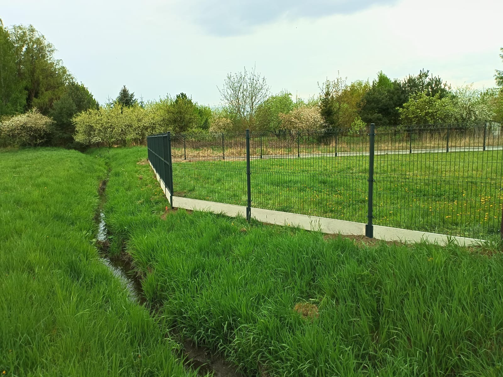Nowoczesne ogrodzenie panelowe w kolorze grafitowym, z podmurówką betonową, oddzielające trawnik od strumyka. W tle zieleń drzew i krzewów.