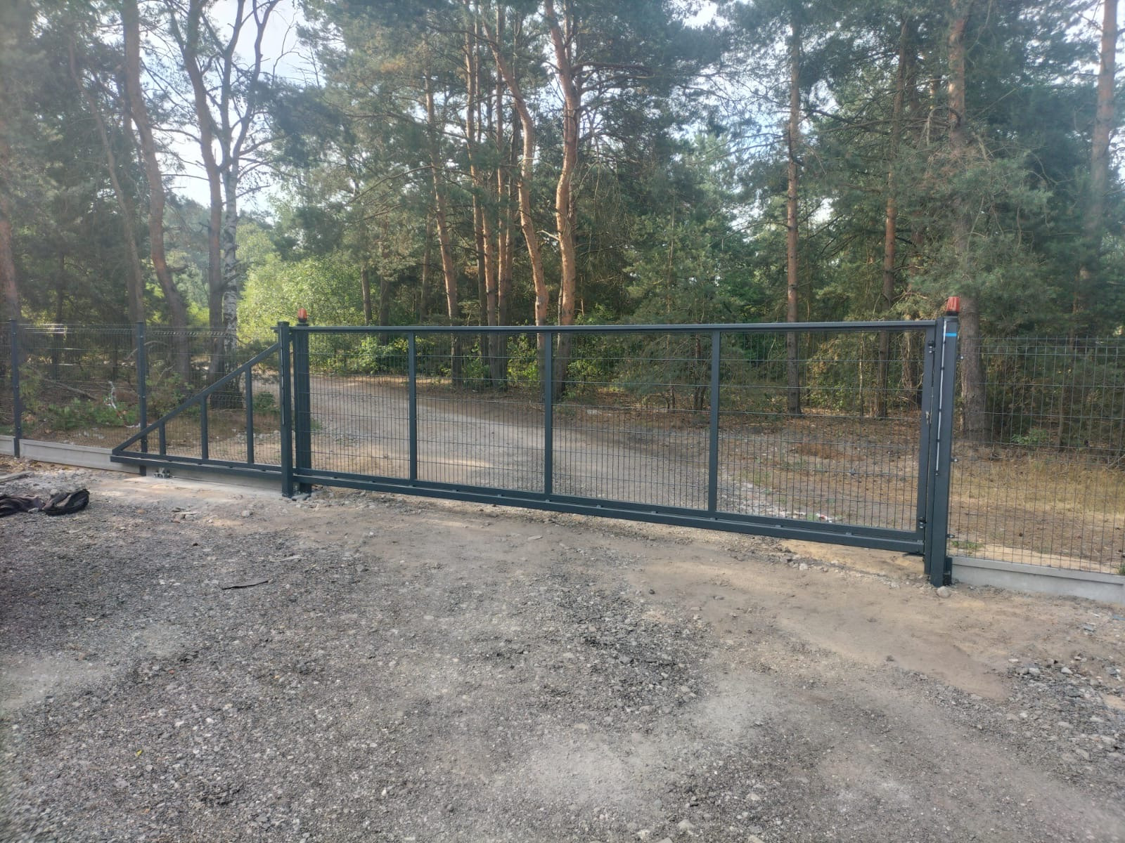 Przesuwna brama z metalowej siatki na tle leśnym. Brama w kolorze grafitowym, zamontowana na betonowej podmurówce. Widoczna linia ogrodzenia z siatki.