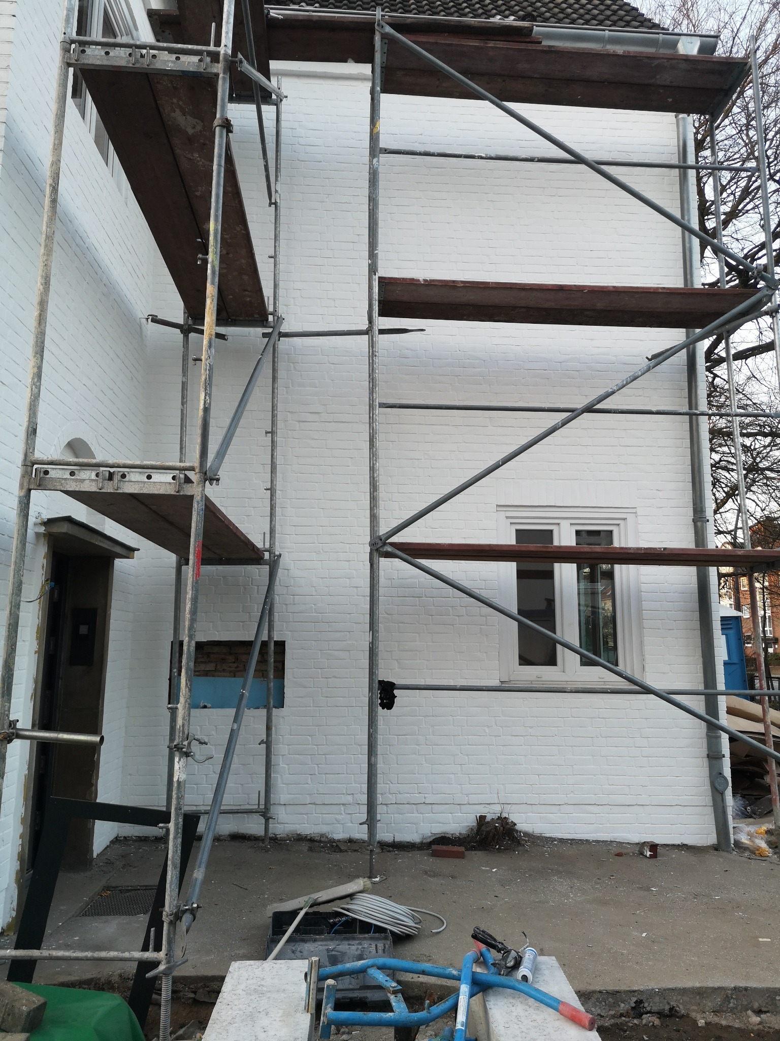 Biała elewacja domu w trakcie prac wykończeniowych, widoczne rusztowania i narzędzia. Świeżo pomalowana ściana z cegły, okno z drewnianą ramą.
