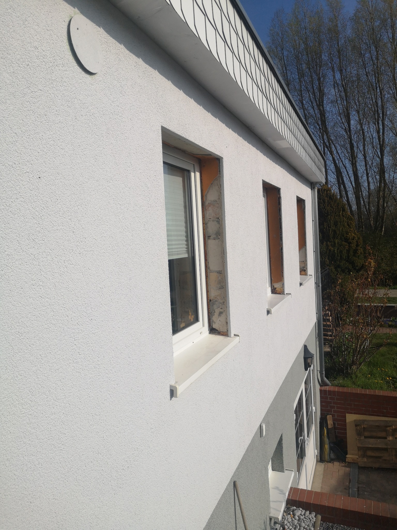 Elewacja domu z oknami PCV w trakcie montażu. Widoczne przygotowane otwory okienne i fragment ocieplonej ściany. Biały tynk strukturalny i szare elementy wykończeniowe.