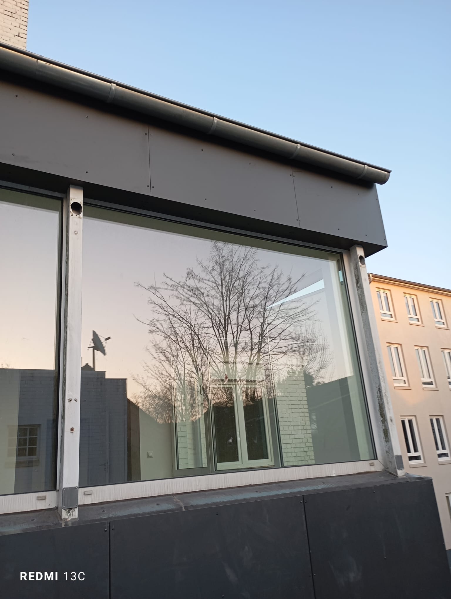 Nowoczesne okno z szarymi ramami, odbijające drzewo i budynki. Widoczne detale montażu i konstrukcji okna, oświetlone popołudniowym światłem.